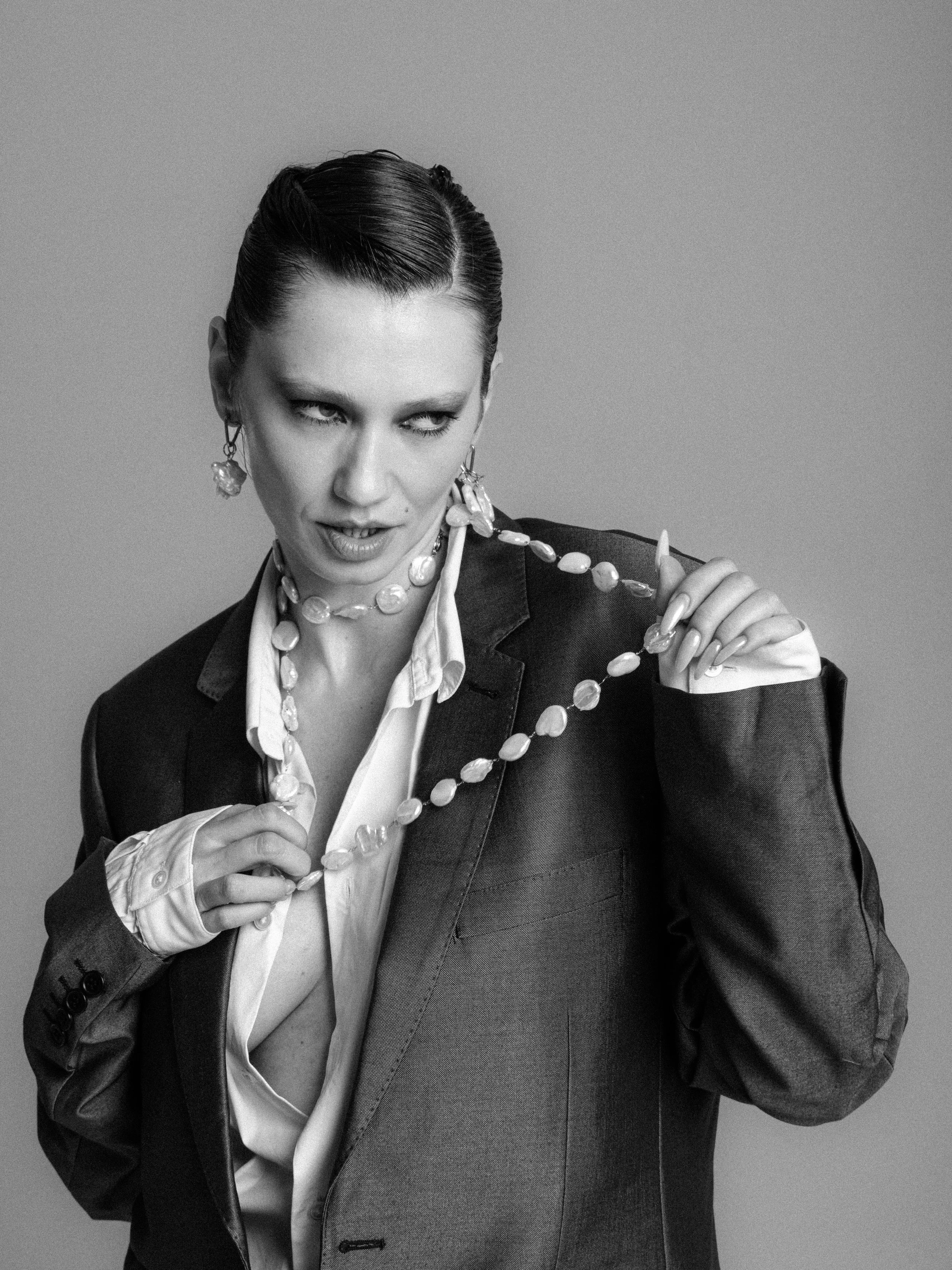 Black and white photo of a woman with slicked-back hair wearing a suit and jewelry, with an intense expression.
