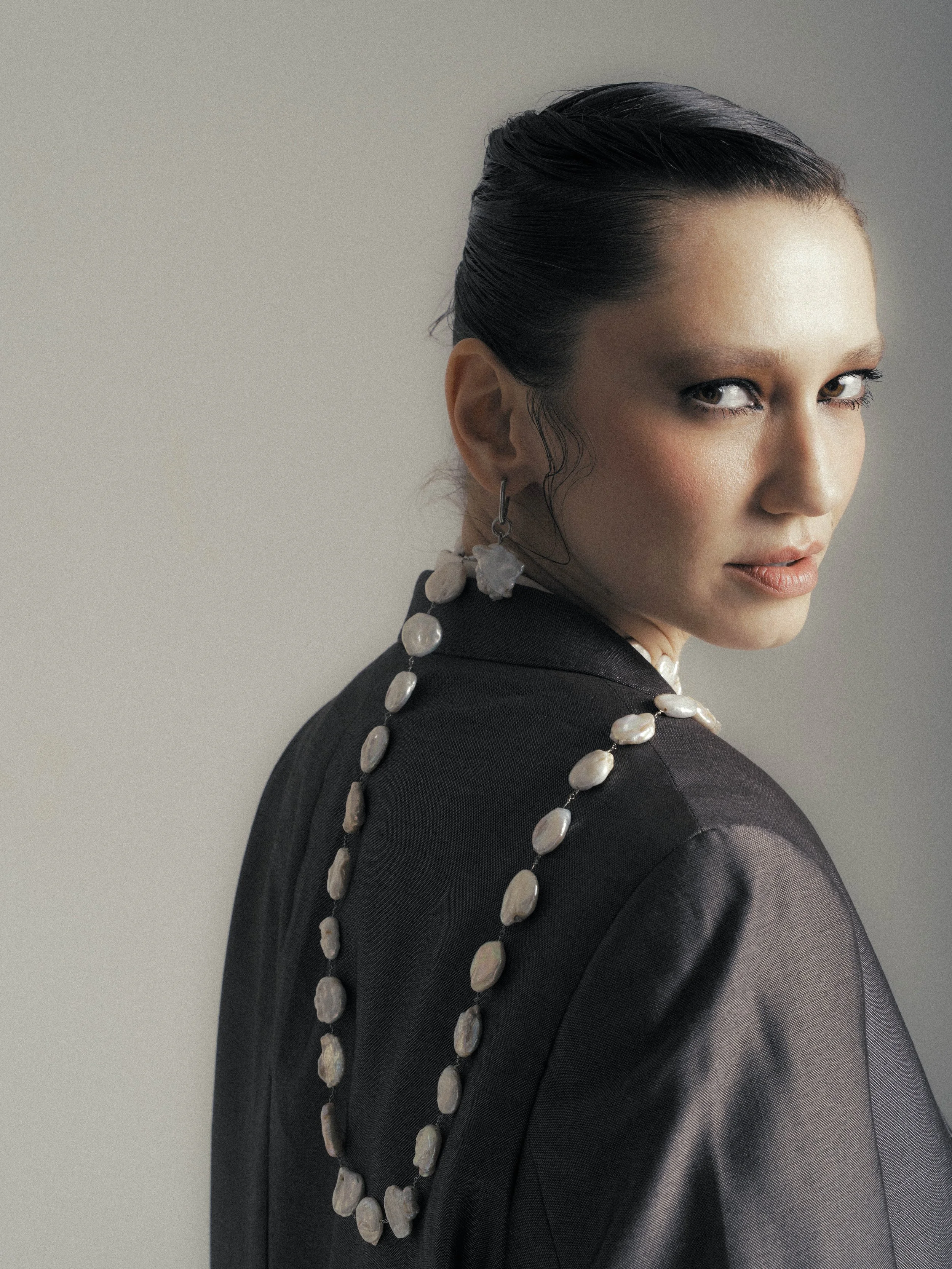 A woman with dark hair styled back, wearing a black blazer, pearl necklace, and matching pearl earrings, looking over her shoulder.