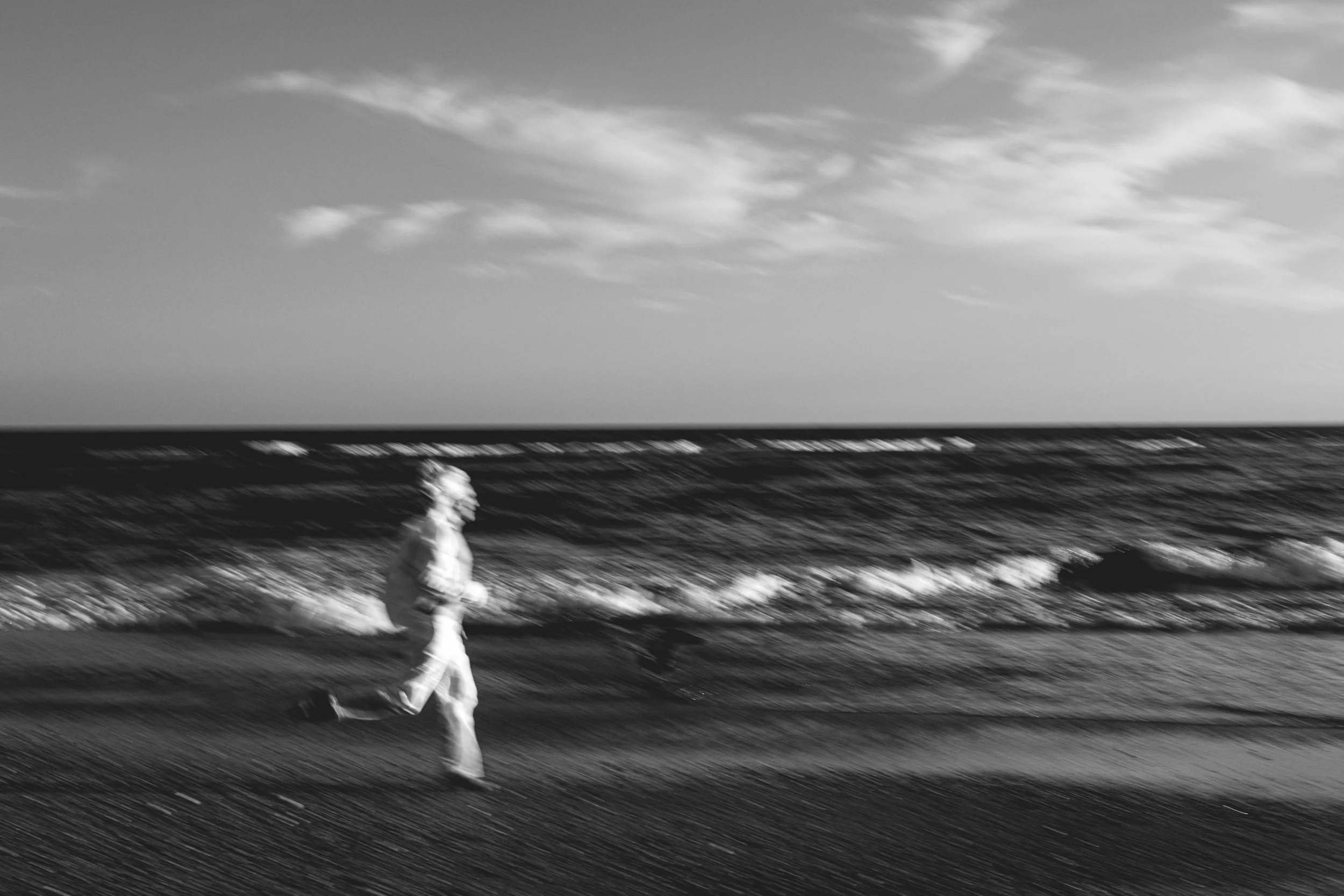 Black and white photo of a person running along the shoreline with the ocean and cloudy sky in the background.