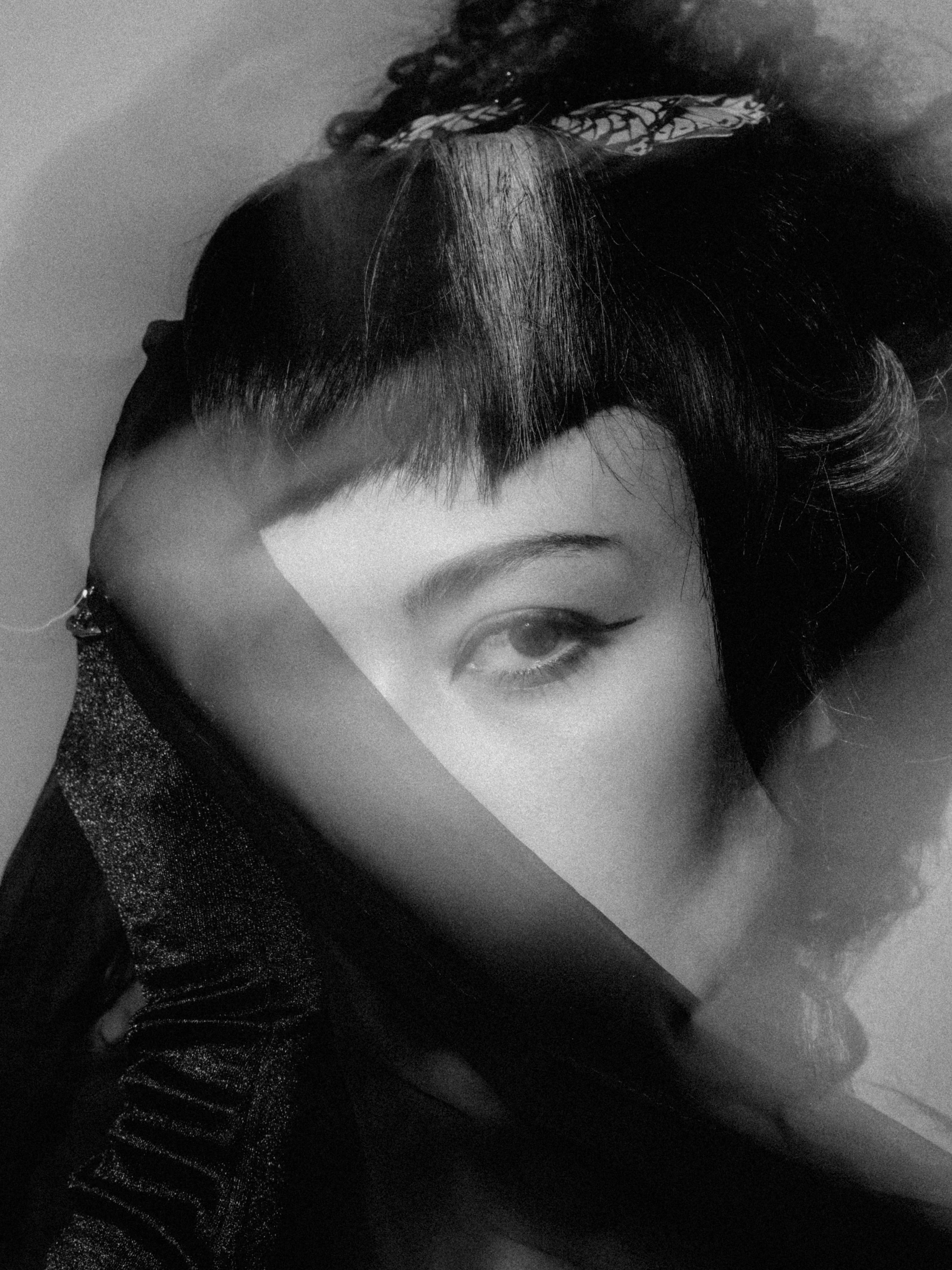 A black and white photo of a woman with dark, wavy hair, styled in a vintage or retro fashion. She is wearing a glittery, shiny top and has a headscarf tied around her head. Part of her face, including her eye and eyebrow, is visible.