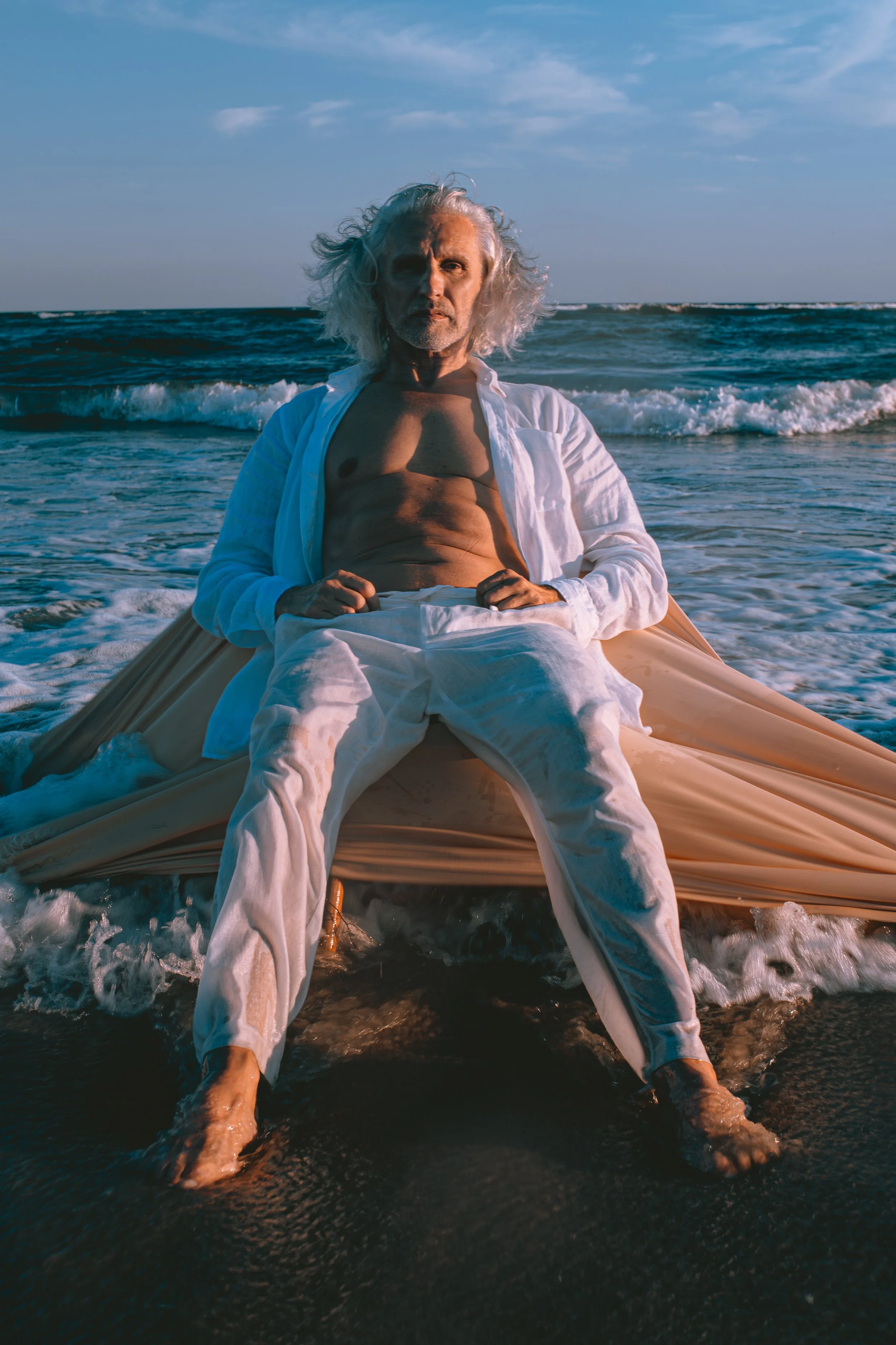 A man with long gray hair and a white shirt open to reveal his chest, sitting on a beach with ocean waves behind him, during sunset or sunrise.