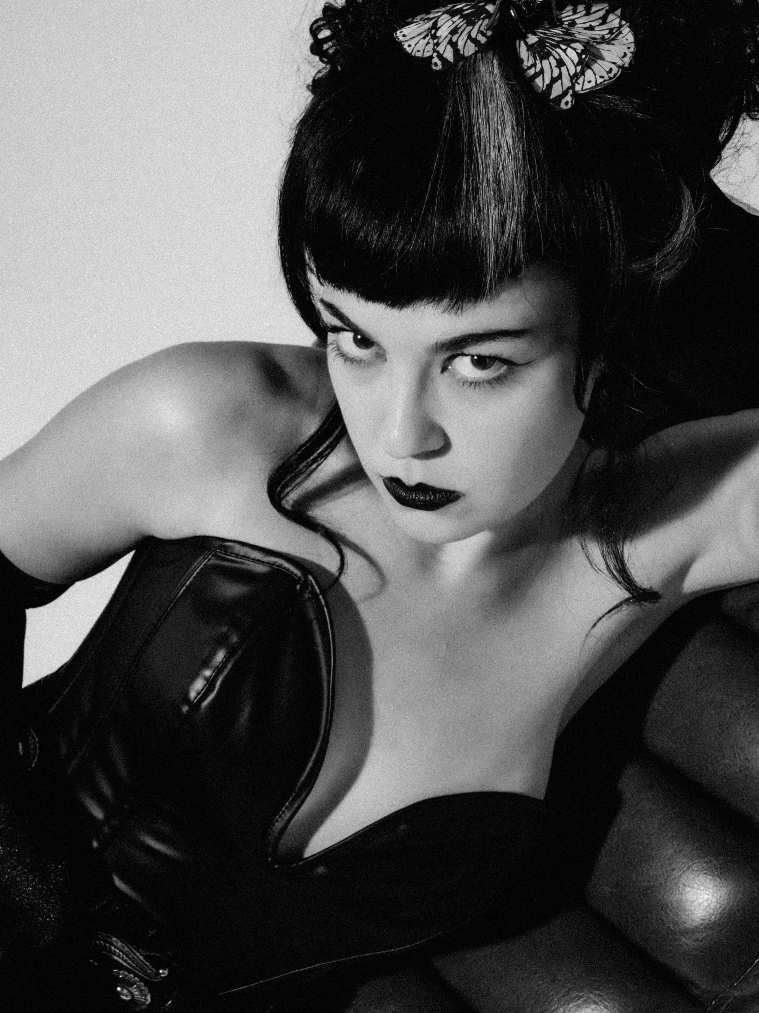 A black-and-white photograph of a woman with dark hair styled in a bob with bangs, wearing a butterfly hair accessory, dark lipstick, and leather clothing, posing in a provocative manner.