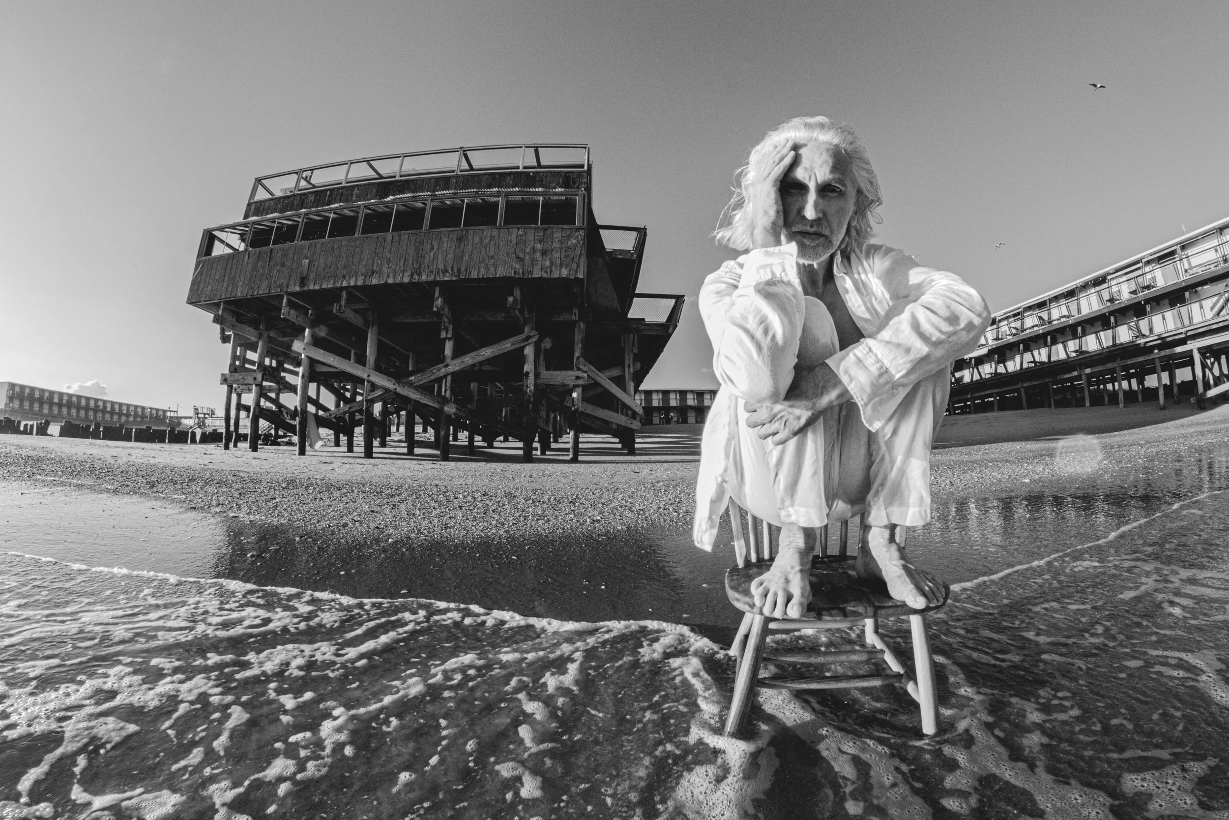 A black-and-white photo of an elderly man with long white hair and a beard sitting on a chair on a beach. He is wearing a light-colored, open shirt and has one hand on his forehead. Behind him, there is a weathered wooden structure on stilts and some