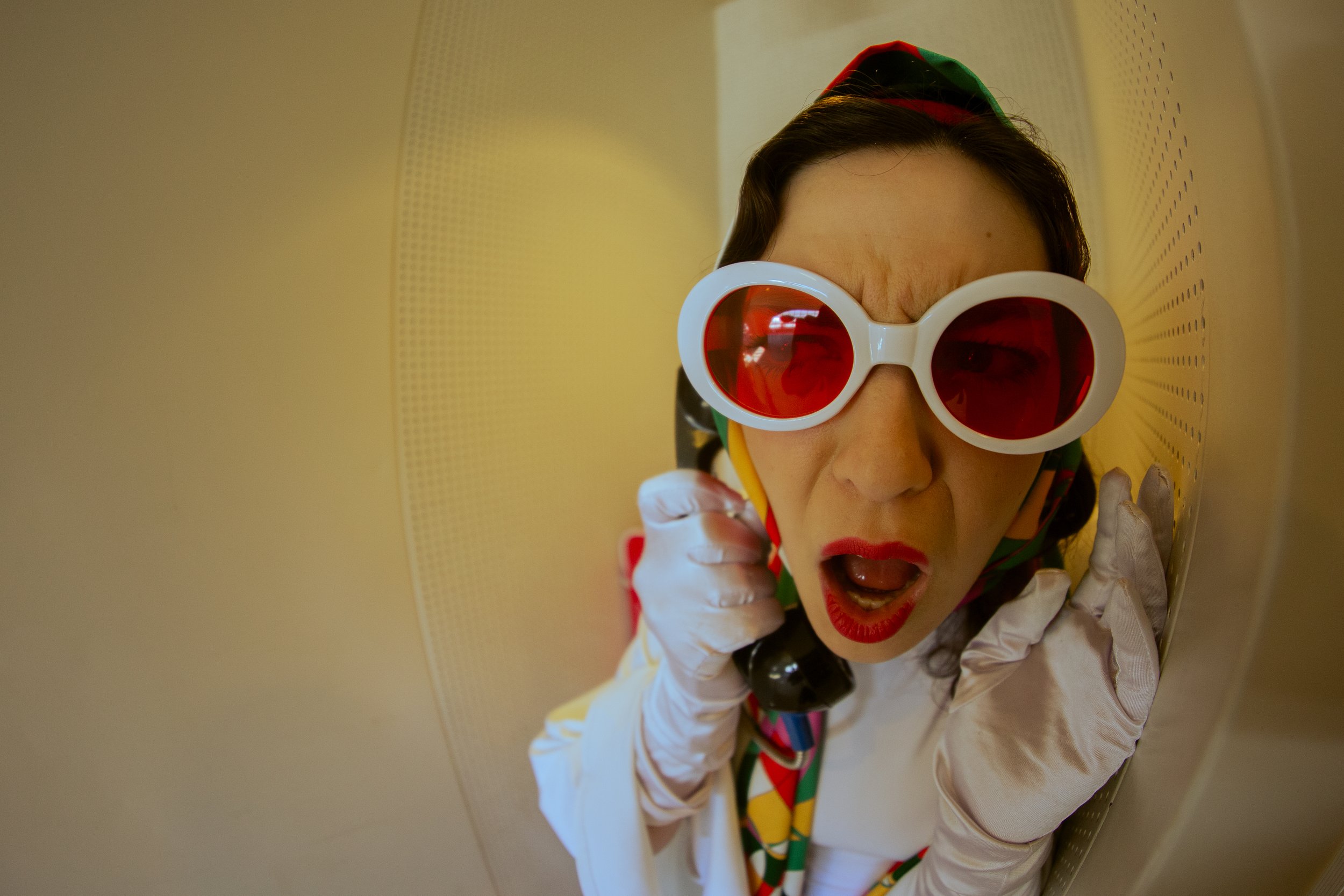 Close-up of a woman wearing large white sunglasses with red lenses, a colorful scarf, and a white outfit, standing against a yellow perforated wall, with a surprised or excited facial expression.