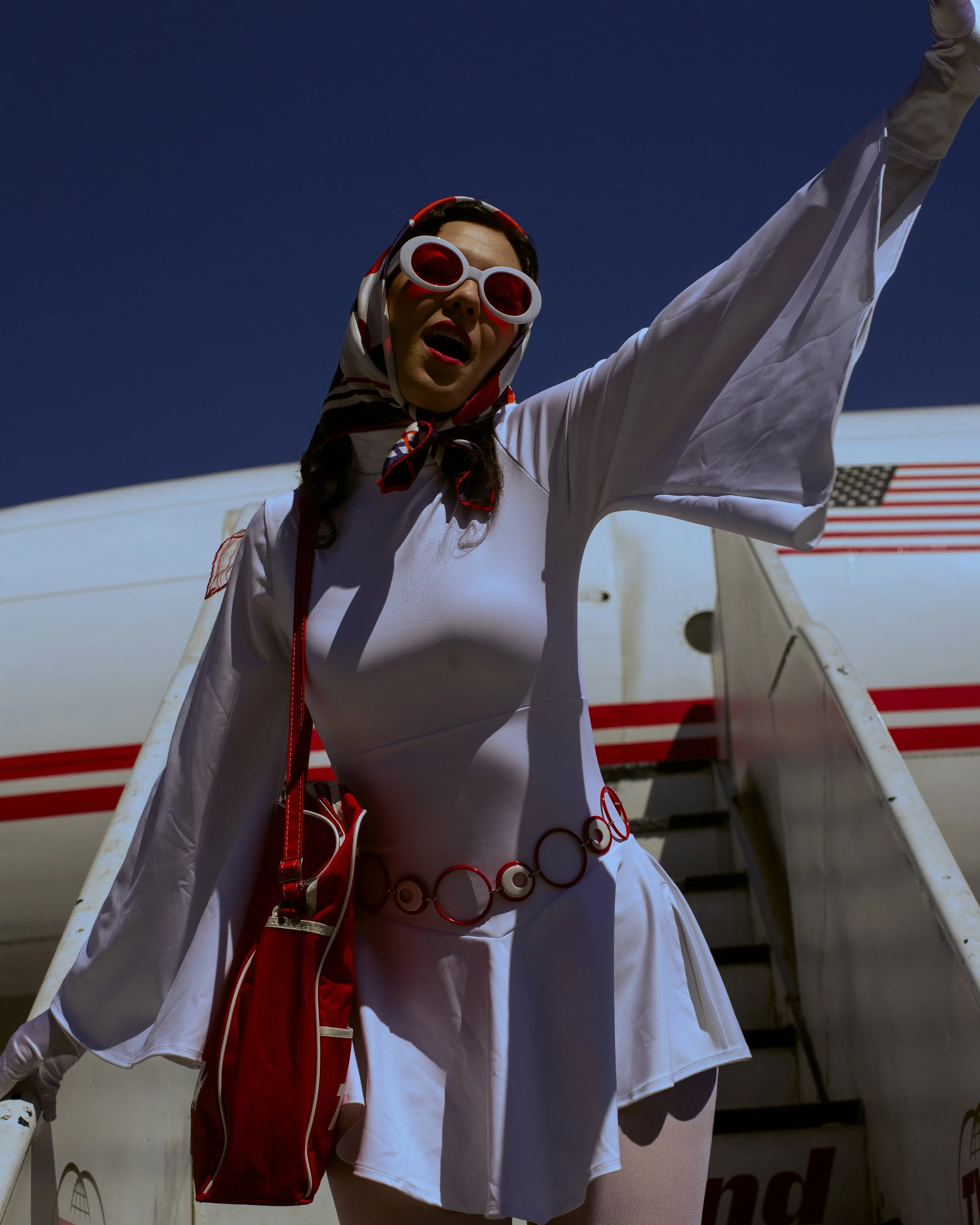 A woman wearing large white sunglasses, a scarf, and a white dress, standing near an airplane with American flag decals.