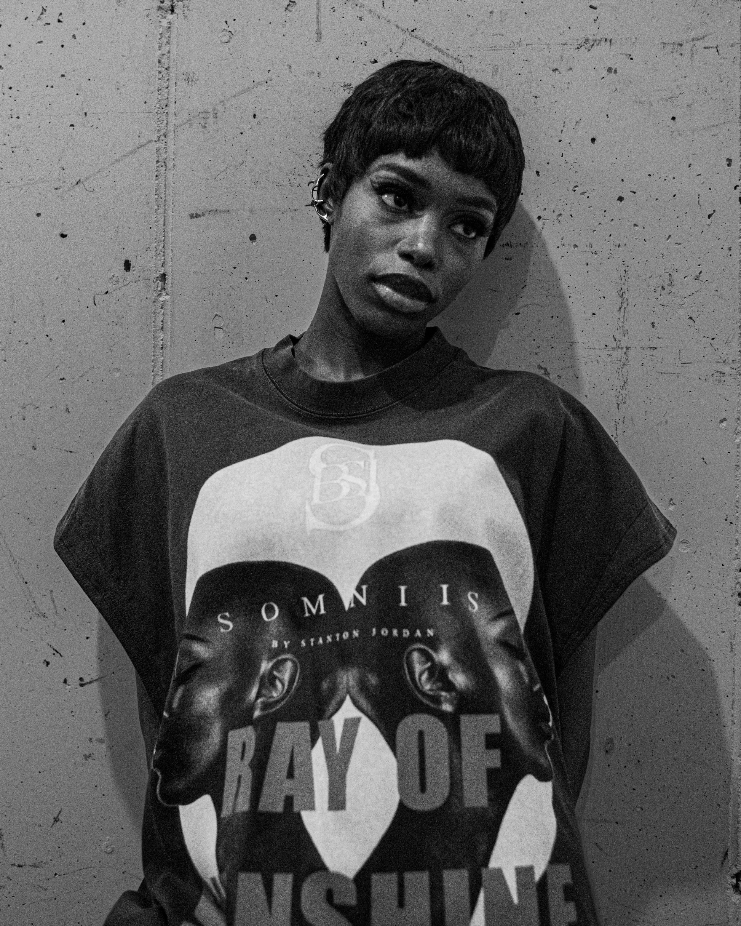 A woman with short hair, dark lipstick, and dark eye makeup is standing against a textured concrete wall. She is wearing a dark T-shirt with a graphic print.