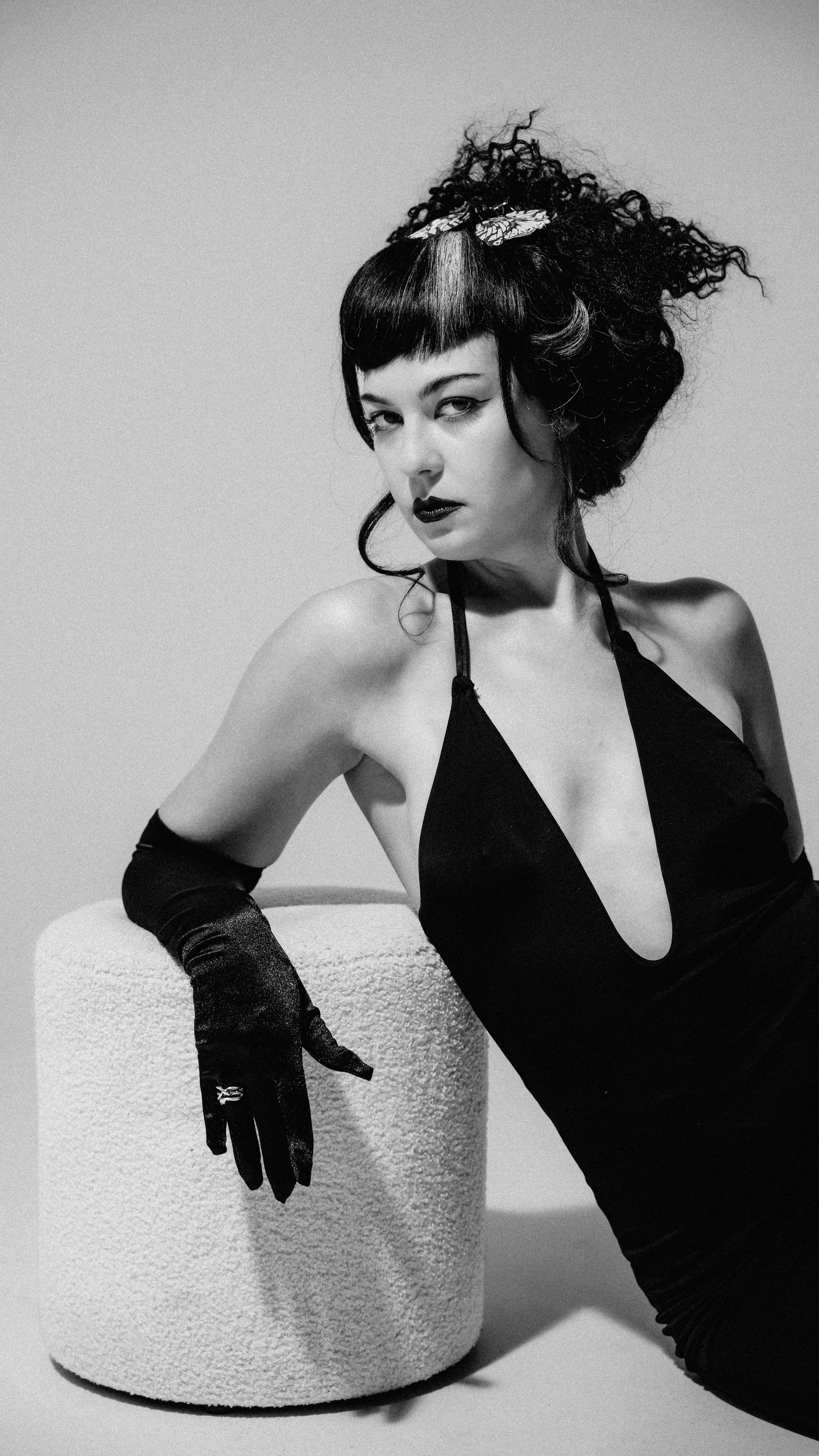A black and white photograph of a woman with short hair, wearing a black dress with a deep neckline, long black gloves, and a decorative headband. She is sitting with one arm resting on a textured cylindrical object while looking directly at the came
