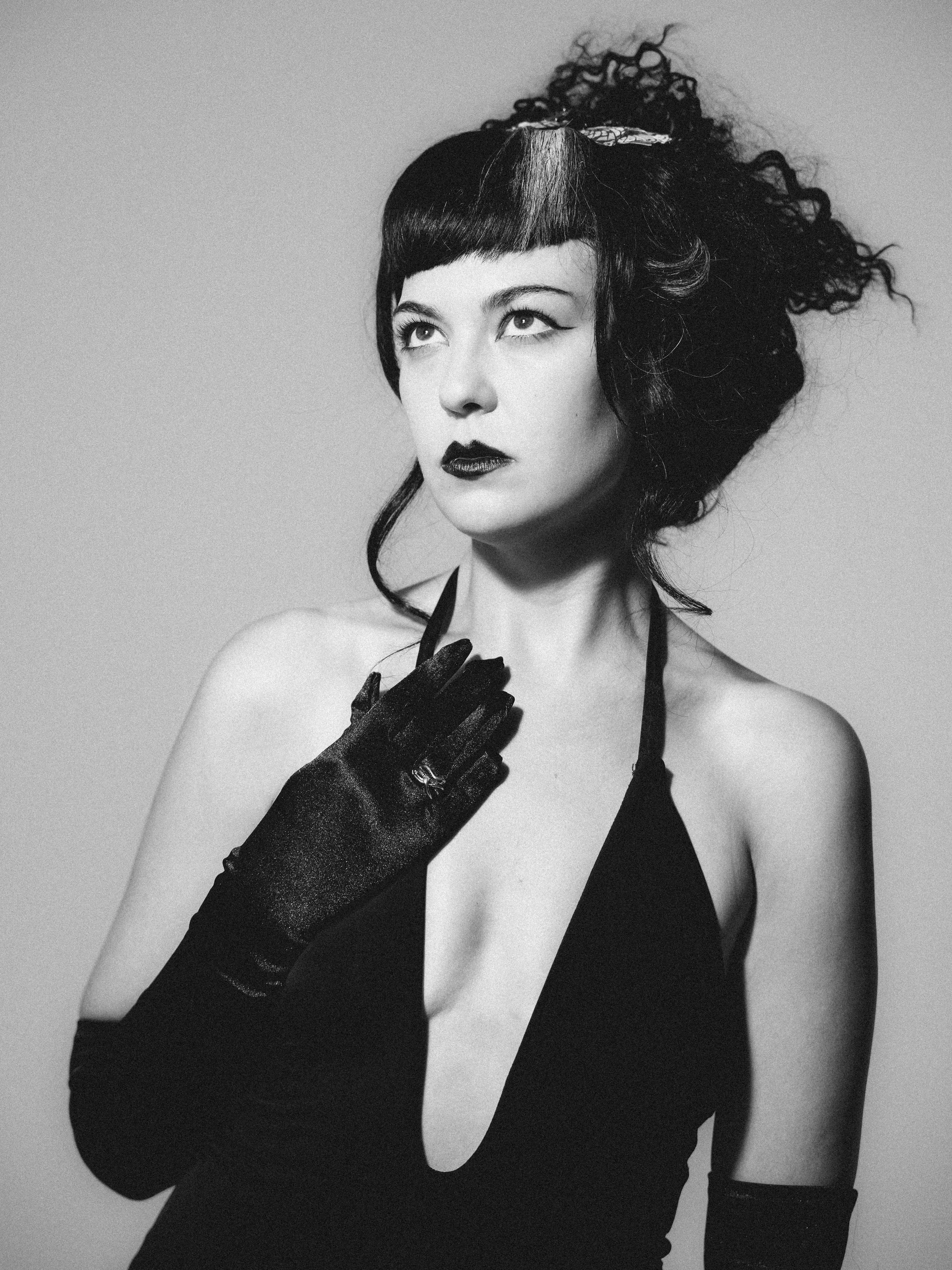 A black and white portrait of a woman wearing a black sleeveless top, black lace gloves, and a black hairstyle with a unique cut and curled details. She has dark lipstick and eye makeup, with her head slightly tilted upward and her gaze directed upwa