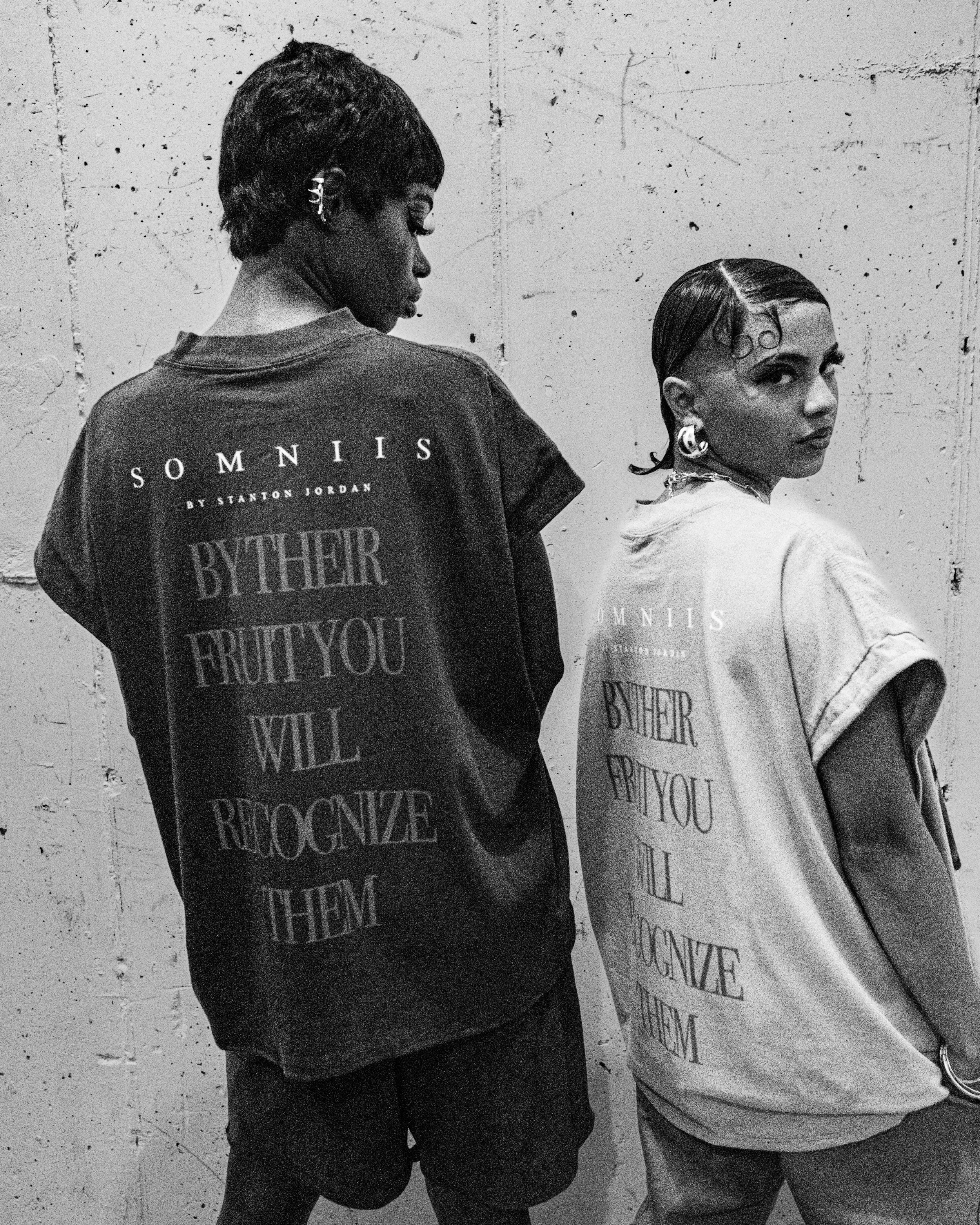 Two women with dark, styled hair and earrings stand against a textured, concrete wall. One woman is facing sideways, wearing a dark t-shirt with text, while the other is facing slightly forward, wearing a light-colored t-shirt with matching text.