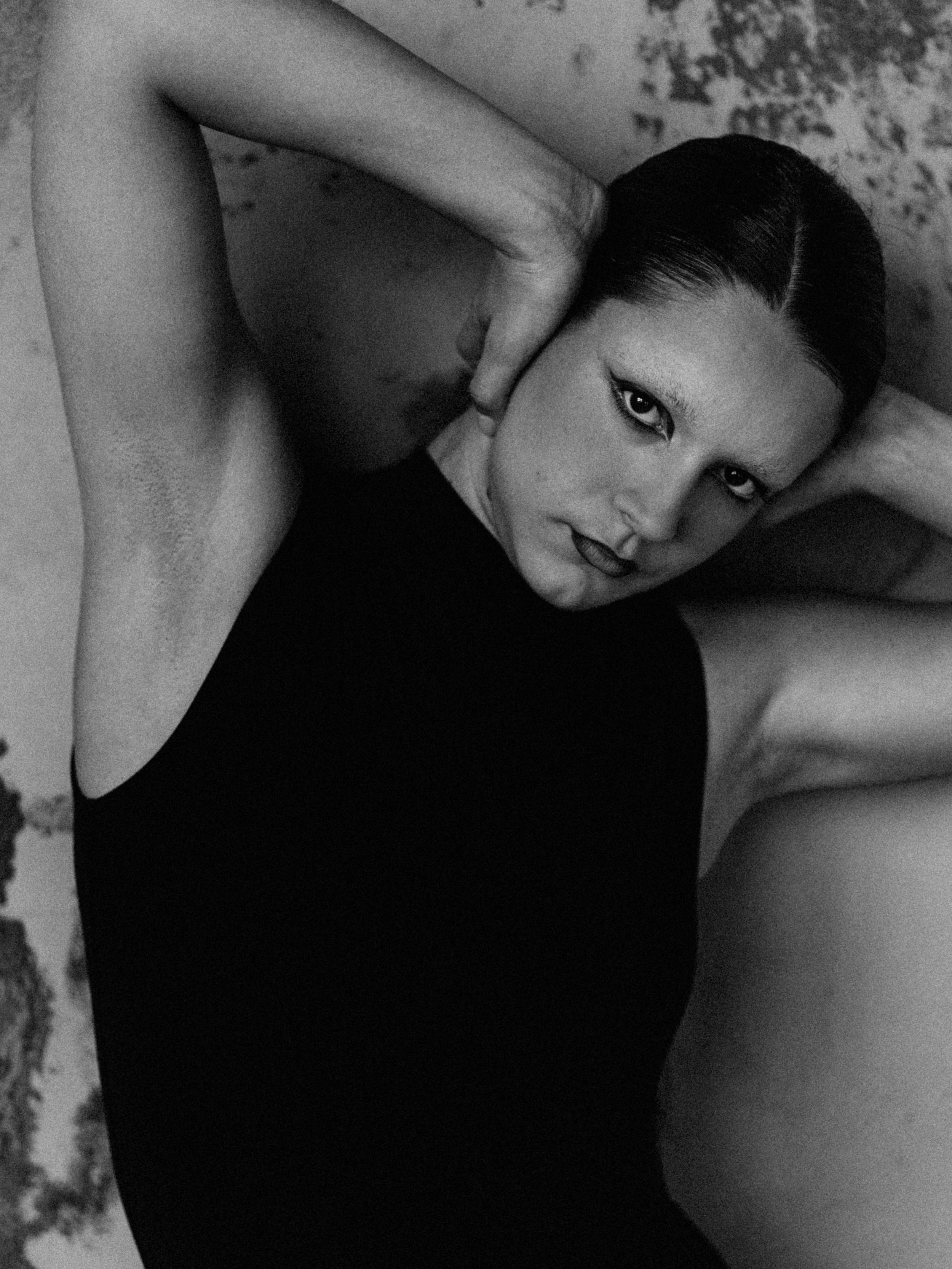 Black and white photograph of a woman with dark hair styled in a sleek updo, lying down with her head tilted and one hand resting on her cheek. She is wearing dark lipstick and a sleeveless top.