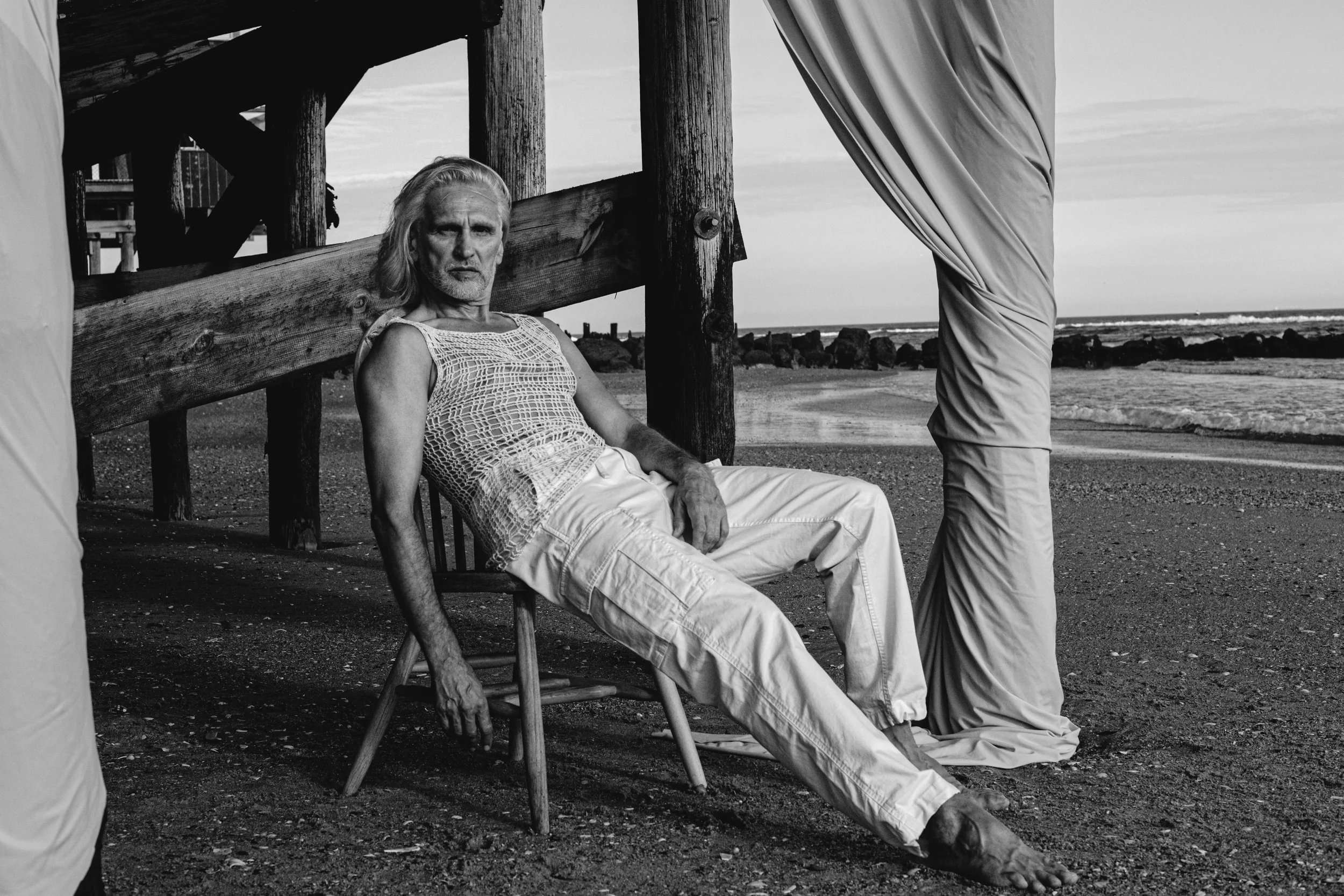 A man with long hair and a beard sitting on a wooden chair on the beach, looking at the camera. There is a person standing nearby, partially visible, and the background shows the ocean and sky.