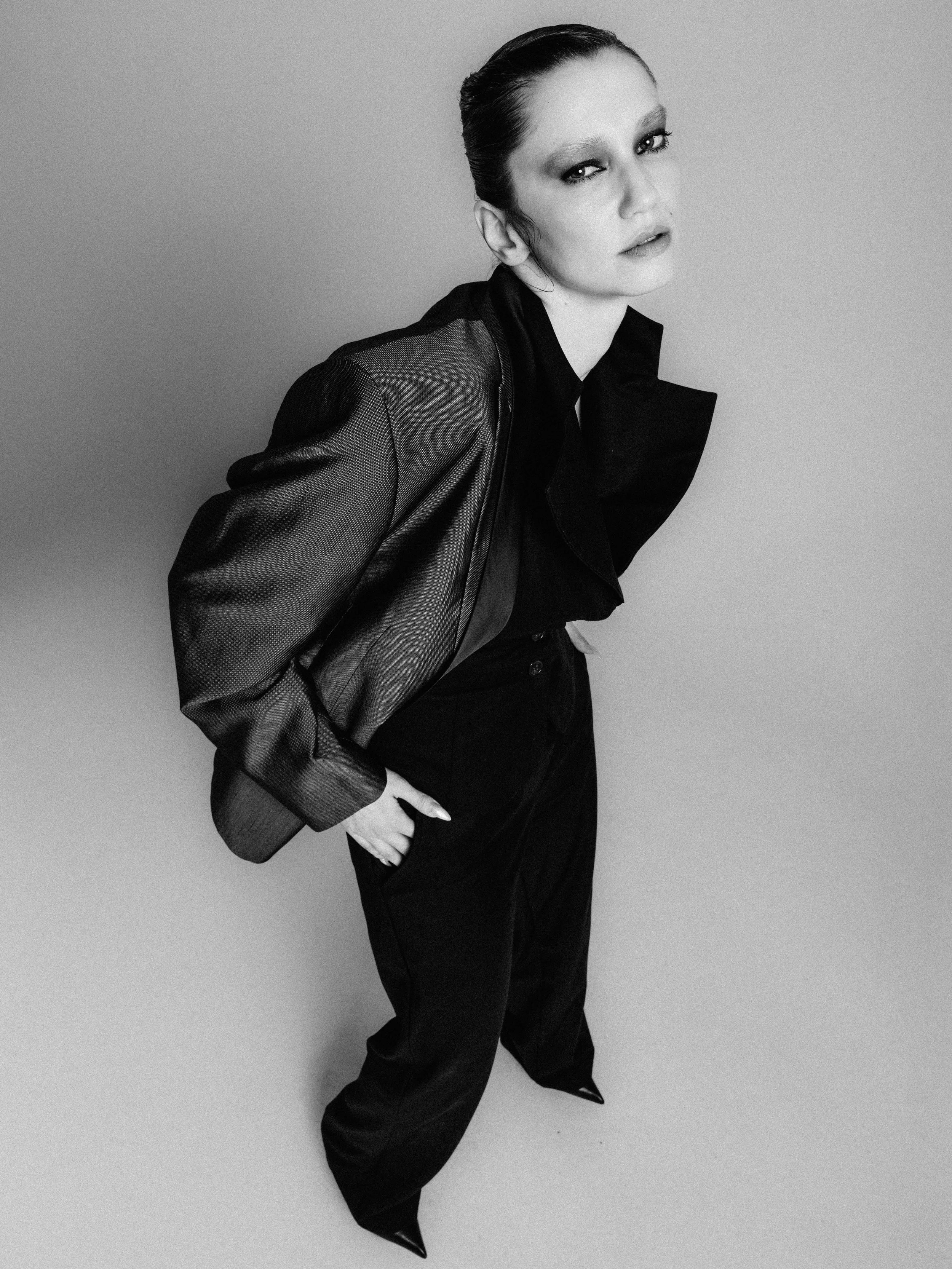 Black and white photo of a woman with slicked-back hair, wearing a shiny, textured jacket and black pants, posing against a plain background.