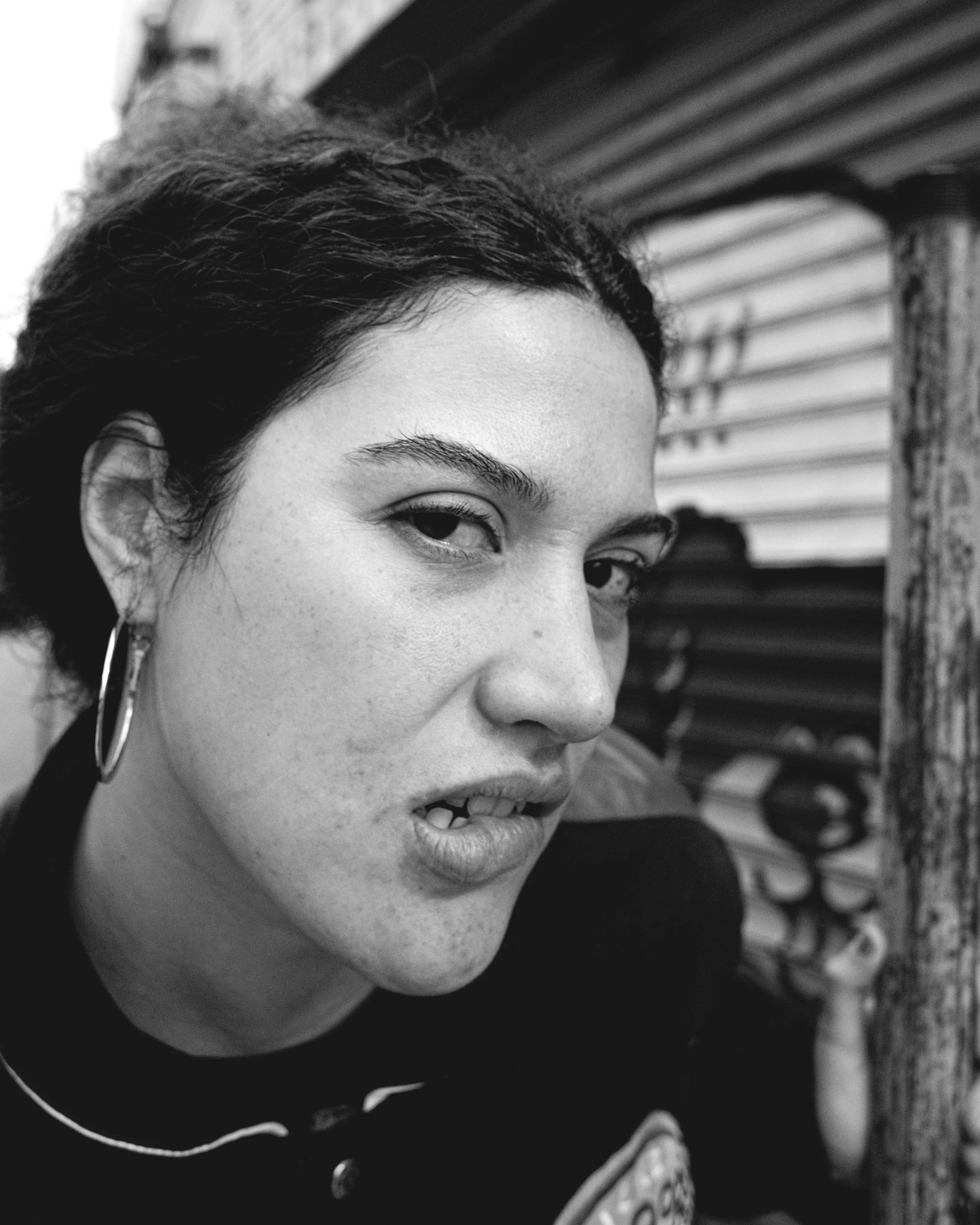 Close-up black-and-white photo of a woman's face, showing her eye, eyebrow, and part of her nose and mouth. She has curly hair, wears large hoop earrings, and appears to be in front of a background with horizontal lines or slats.