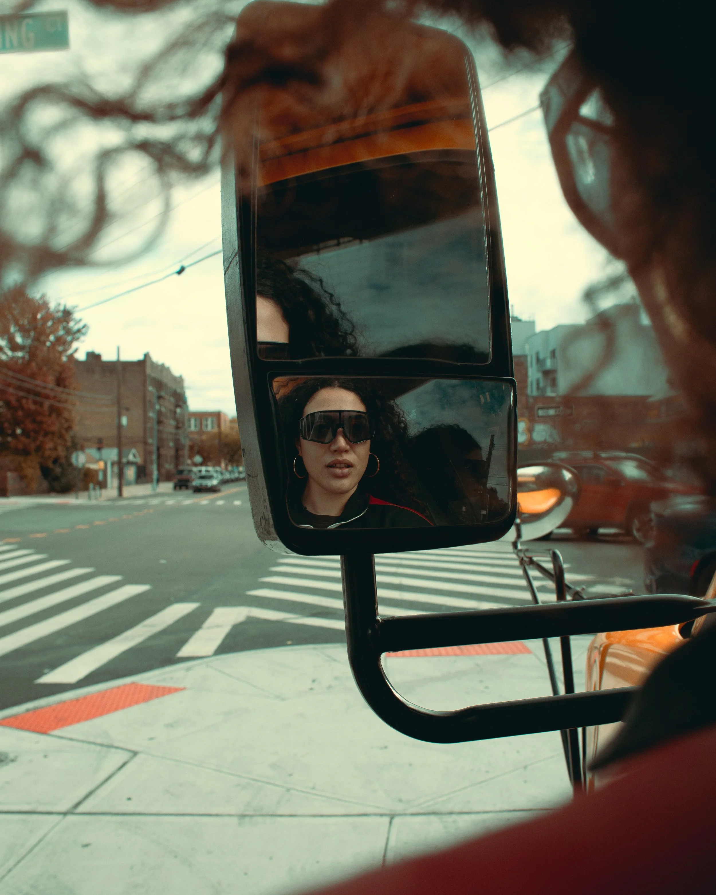 Reflection of a woman with curly hair and sunglasses in a vehicle rearview mirror, with city street scene including crosswalk and buildings in the background.