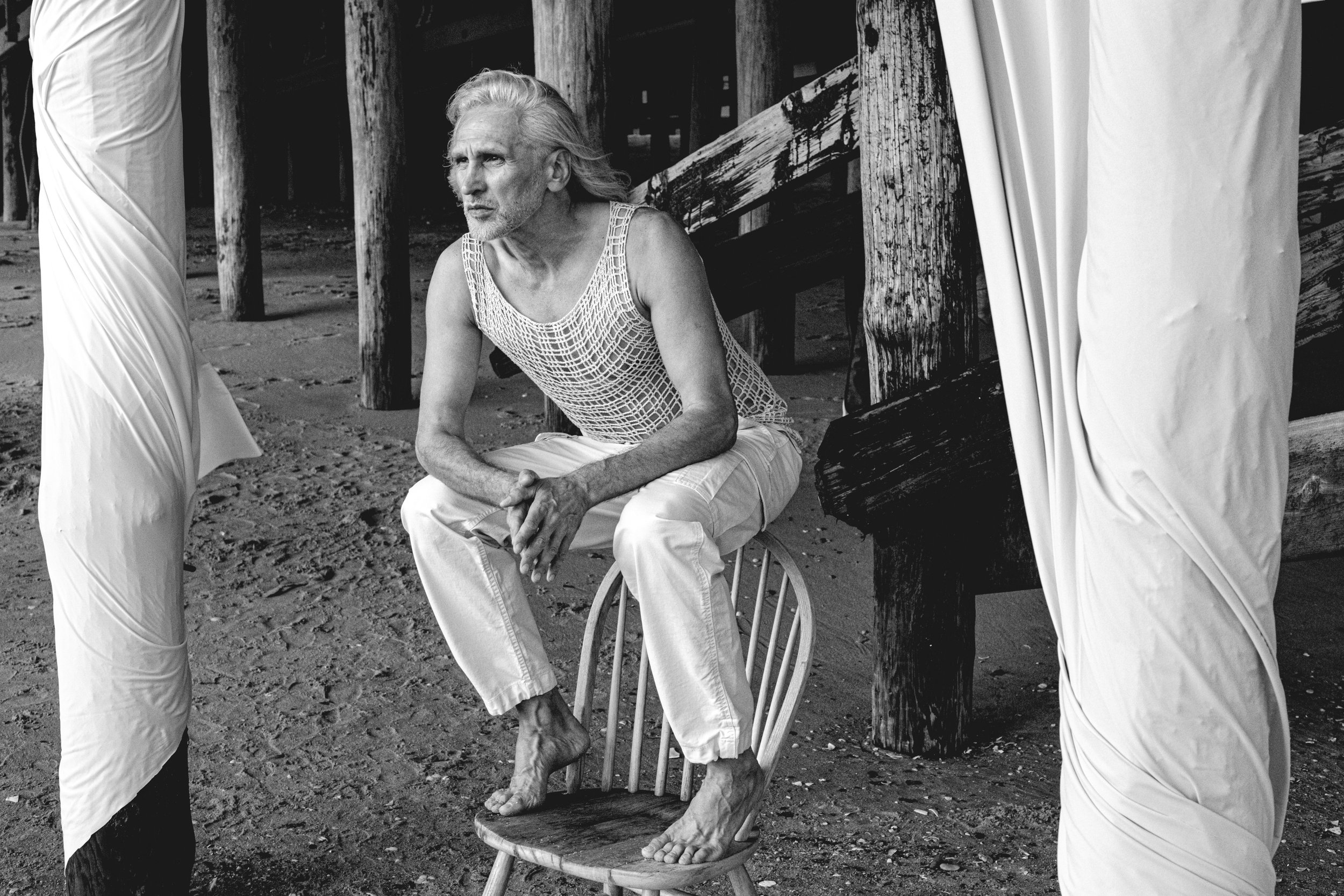 An elderly man with long gray hair and a beard, sitting on a stool with his feet on the ground, on a beach near wooden pilings. He is wearing a crochet tank top and light-colored pants, with a serious expression. The scene is in black and white.