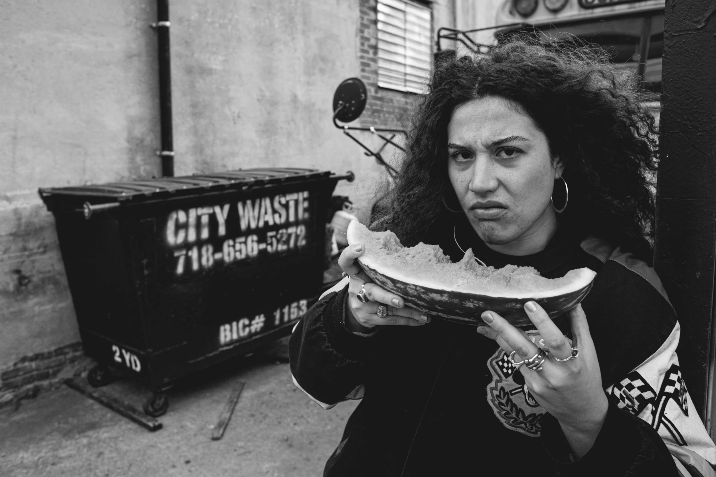 A woman with curly hair holding a large slice of watermelon, standing next to a black city waste bin outdoors.