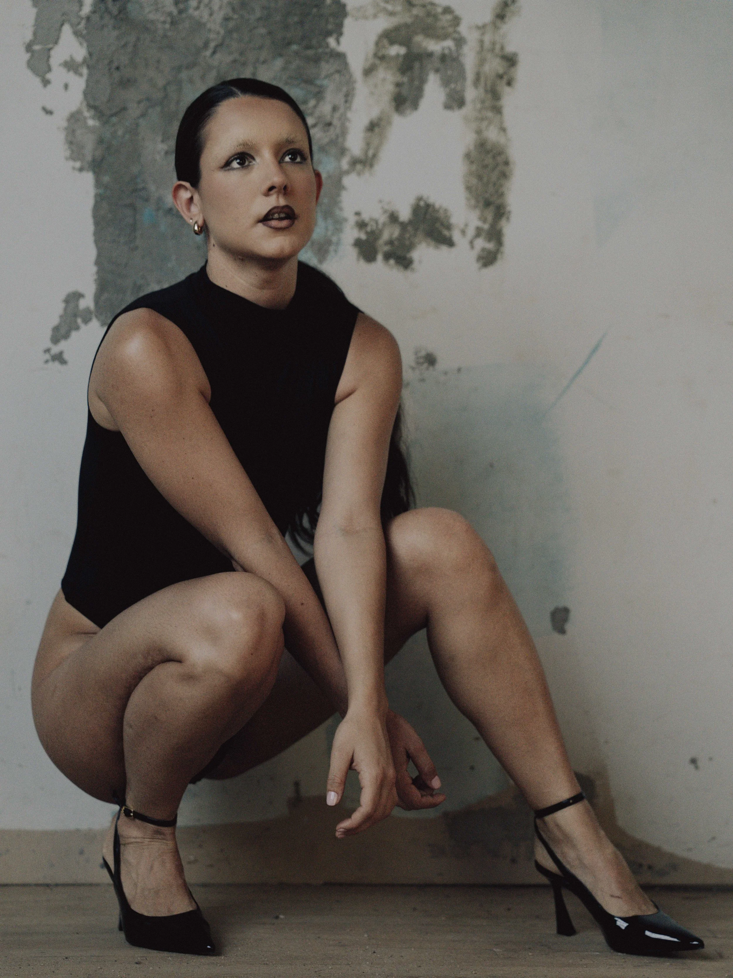 A person with short dark hair and dark makeup, wearing a black sleeveless top, black high heels, and sitting on the floor with crossed legs in front of a distressed wall with peeling paint and a partial map of the world.
