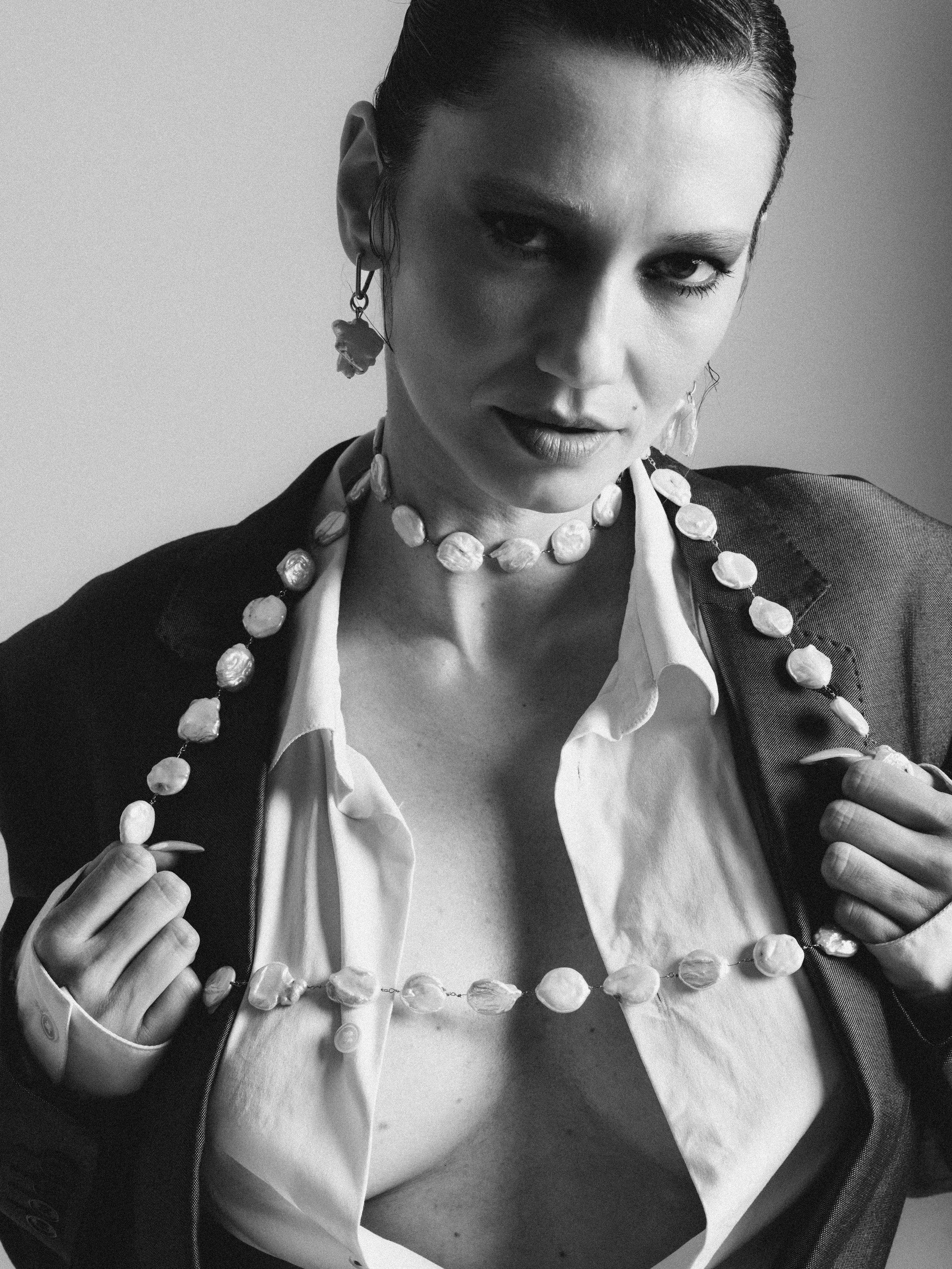 A woman wearing a white blouse and a black jacket with a necklace and earrings, adjusting her necklace with both hands.