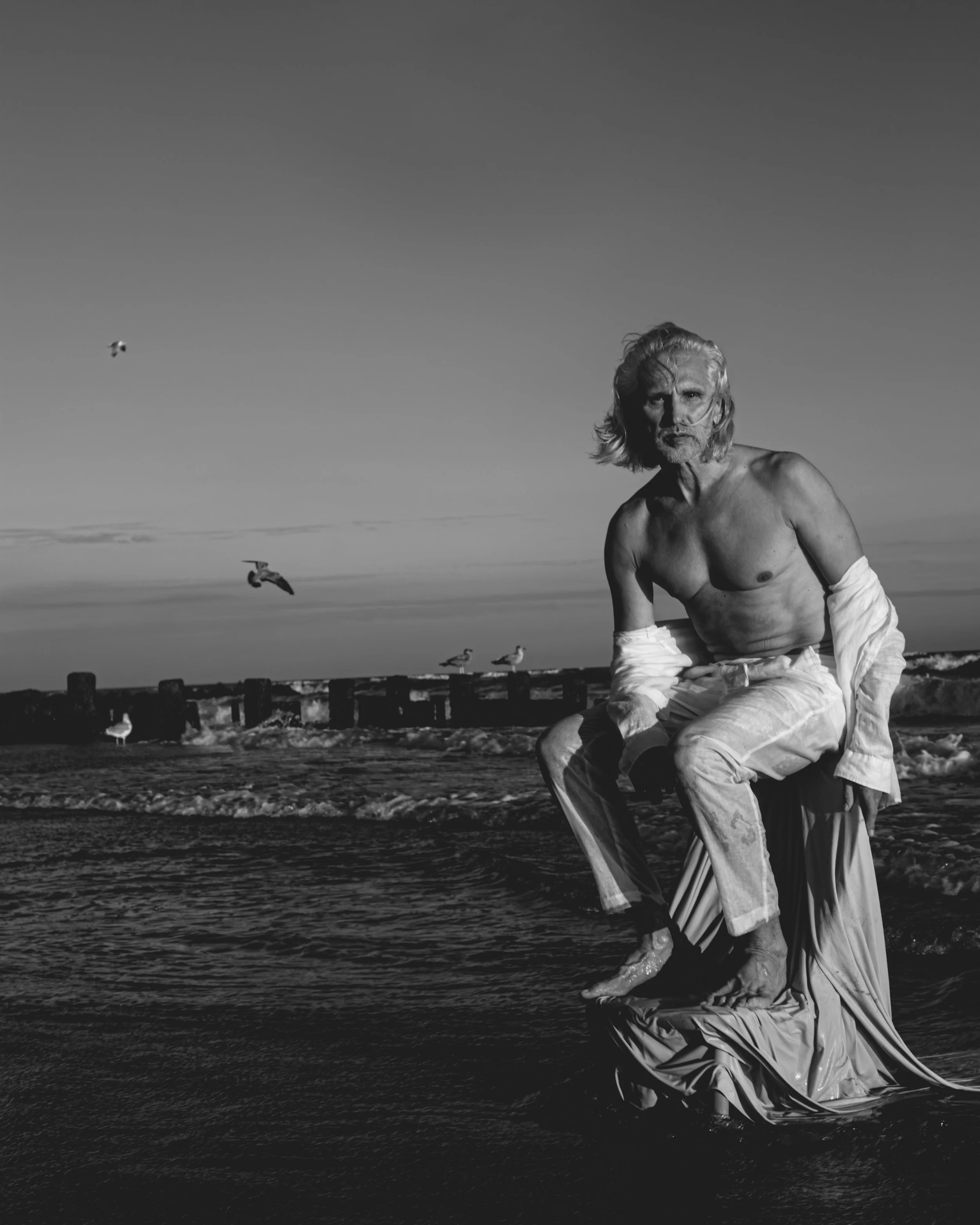 A black and white photo of a shirtless man with long, wavy hair and a beard sitting on a rock at the beach. Seagulls are flying in the sky, and a wooden pier extends into the ocean in the background.