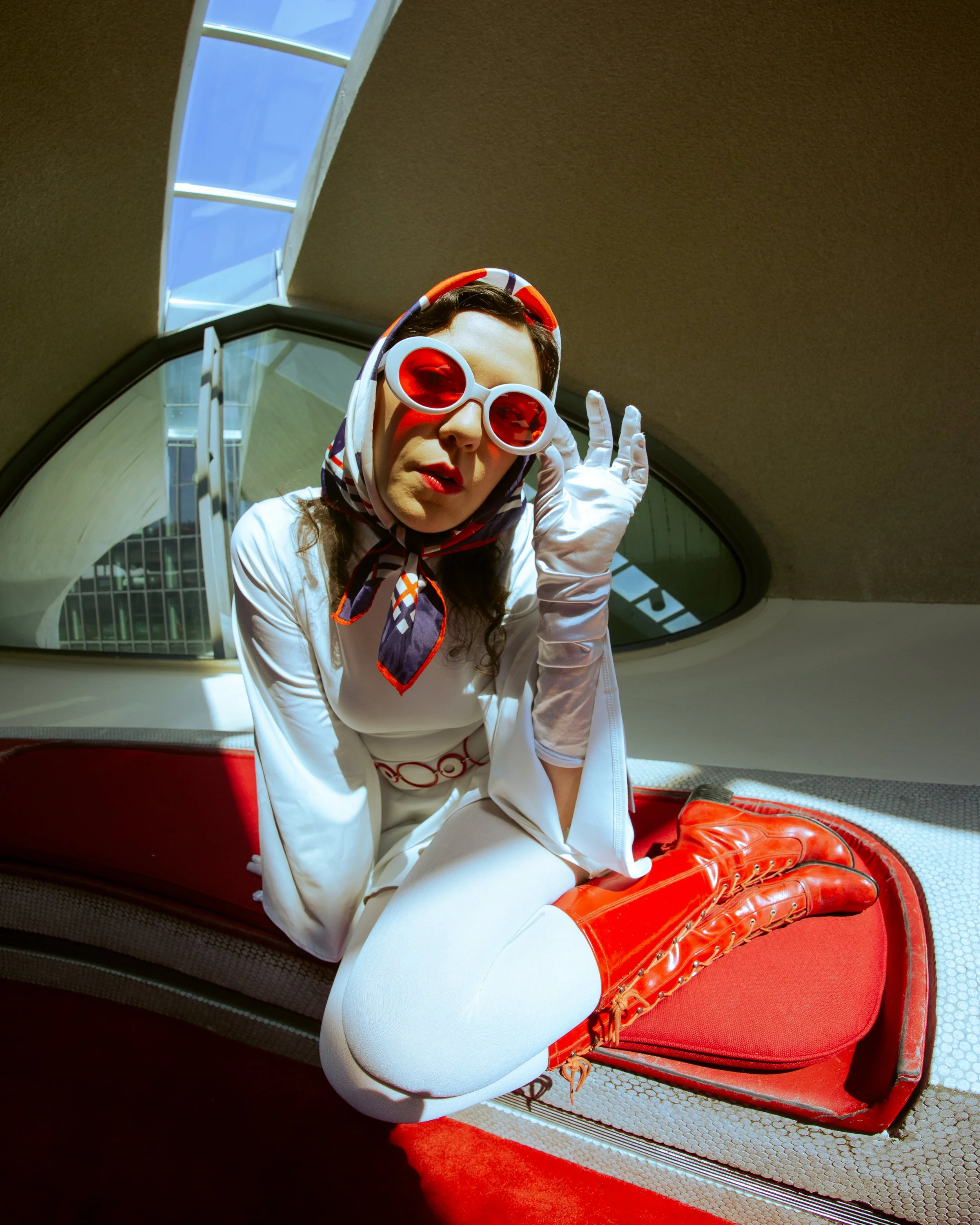 A woman wearing large red sunglasses, a poppy headscarf, and red boots, sitting on the floor with her legs bent, inside a modern building with curved architecture and large windows.