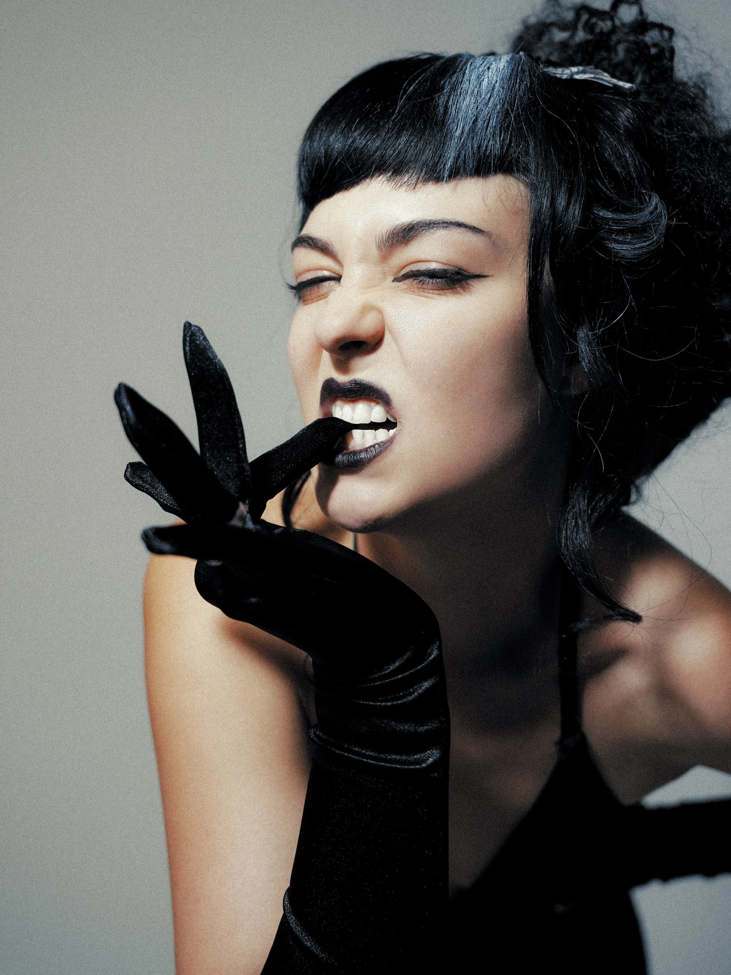 A woman with dark hair styled in a vintage look, wearing black gloves, bold makeup, and biting her finger.