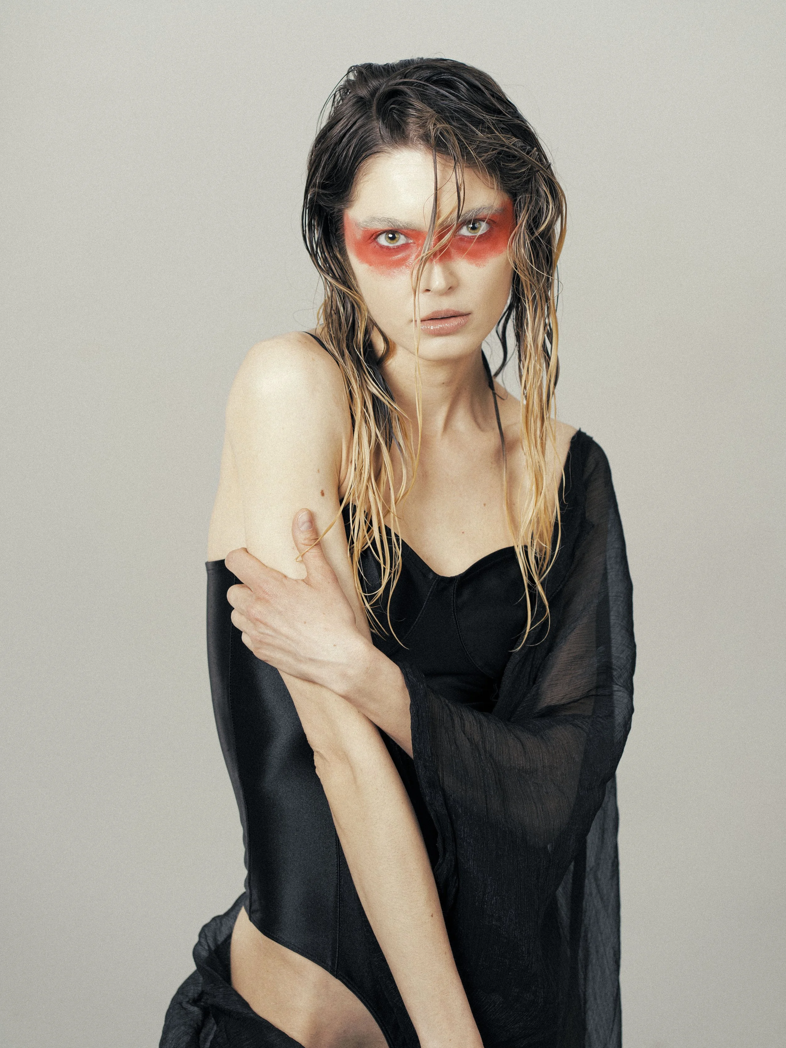 A woman with wet hair and pale skin, wearing a black sheer top, has red eyeshadow around her eyes, creating a bold makeup look. She looks directly at the camera with an intense expression, with her arms crossed in front of her.