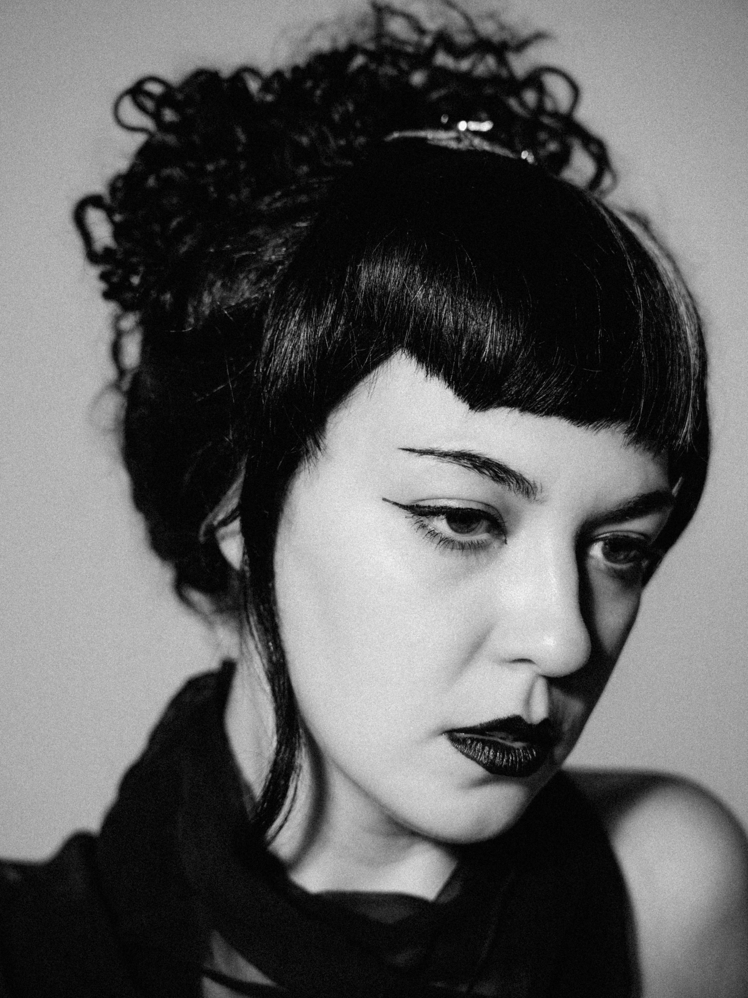 A black and white close-up of a woman's face showing her eyes, nose, lips, and part of her black clothing. She has dark, wavy hair styled with a bow, and makeup with dark lipstick and eyeliner.