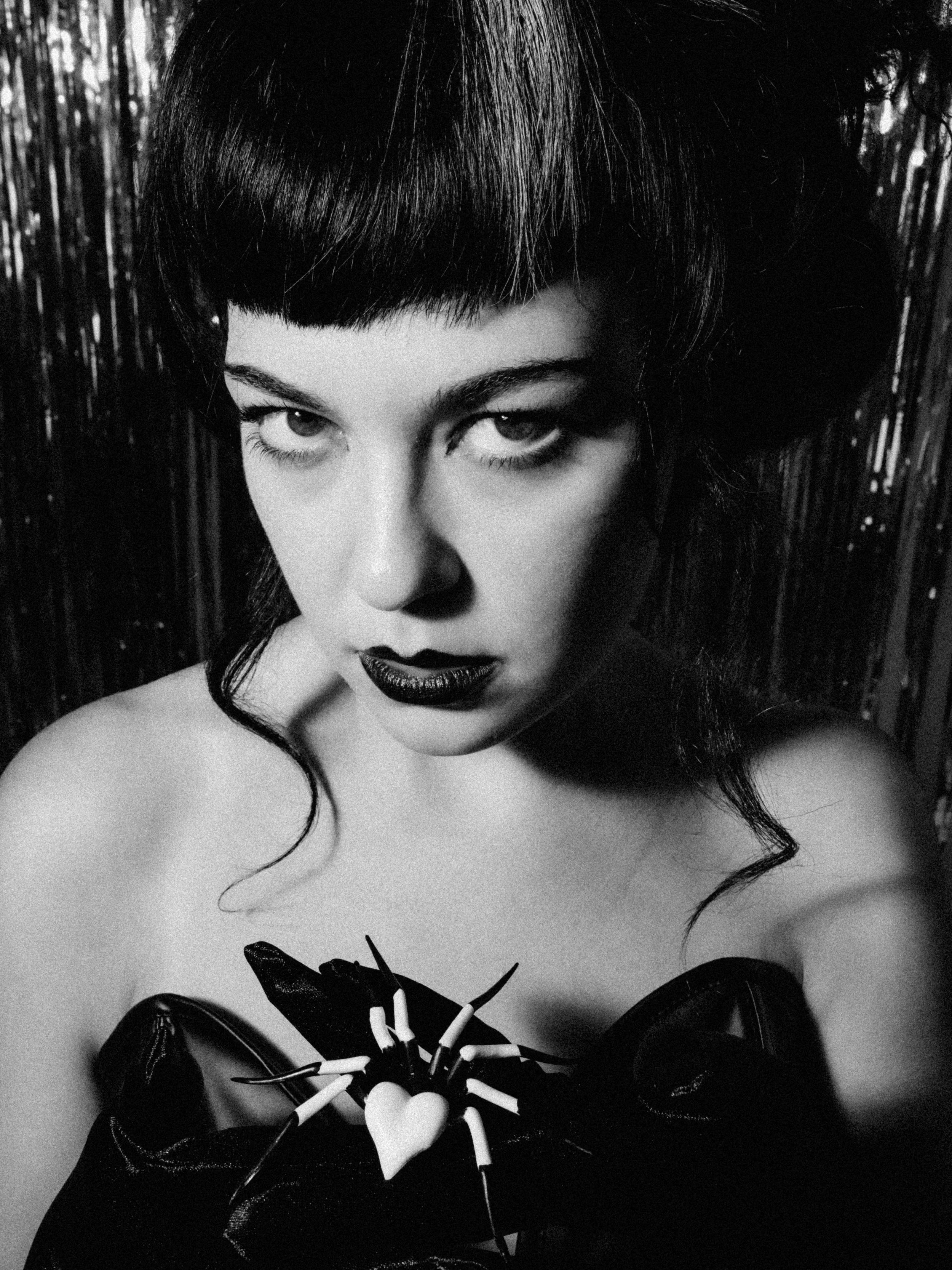 Black and white close-up photo of a woman with dark hair, styled with bangs and voluminous hair, wearing dark lipstick and dark clothing, holding a black heart-shaped object with pale tentacle-like sticks, against a dark textured background.