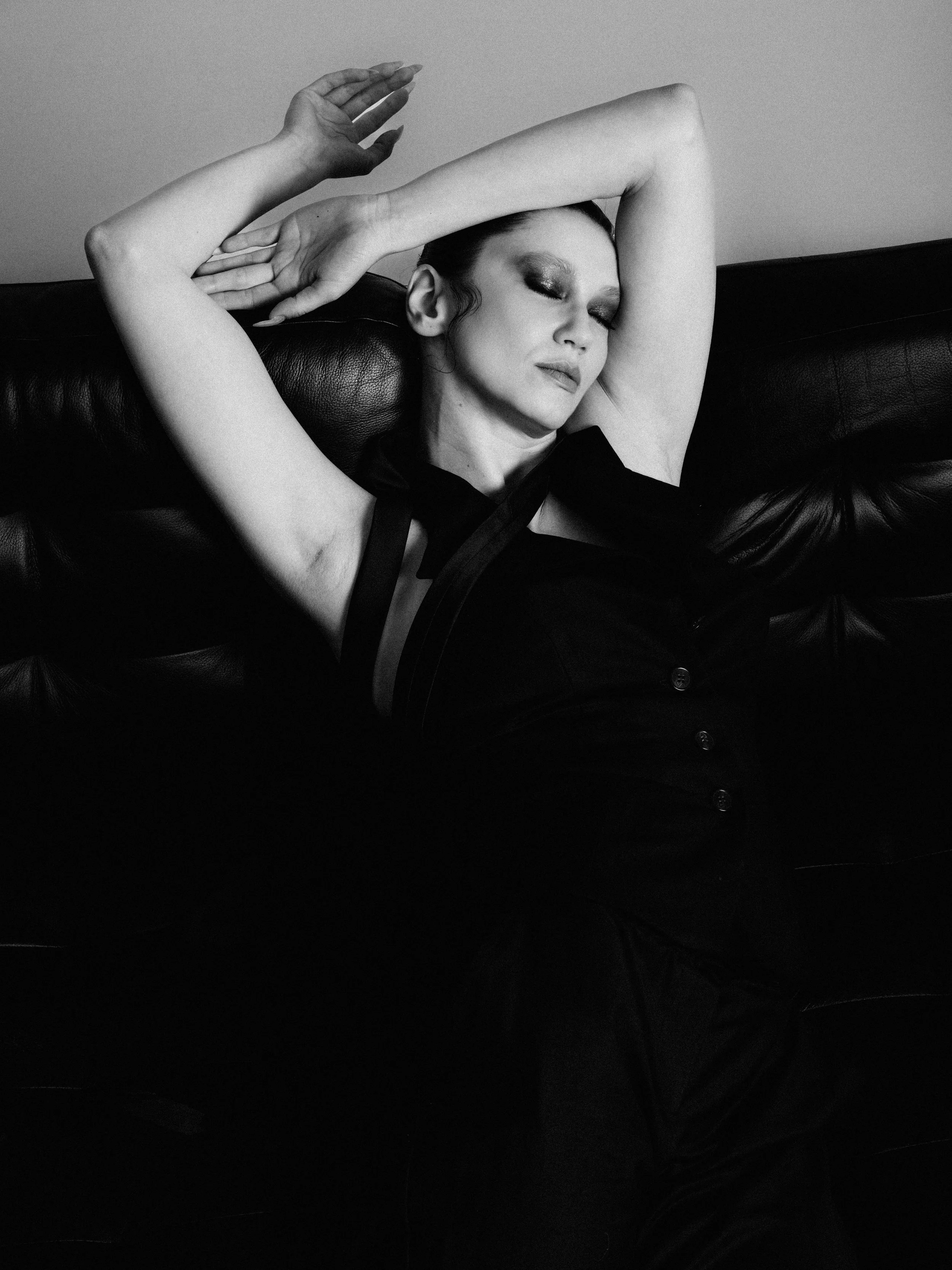Black and white photo of a woman with her eyes closed, head tilted back, and arms raised above her head, sitting on a black leather couch.