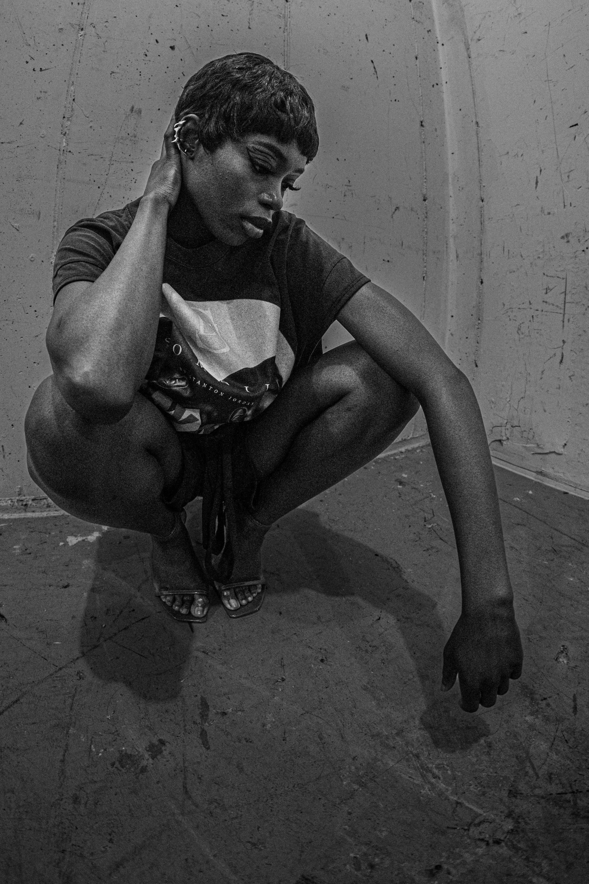A person with short hair and multiple earrings, wearing a T-shirt, squatting down and touching their head, in an industrial setting with a rough, textured wall.