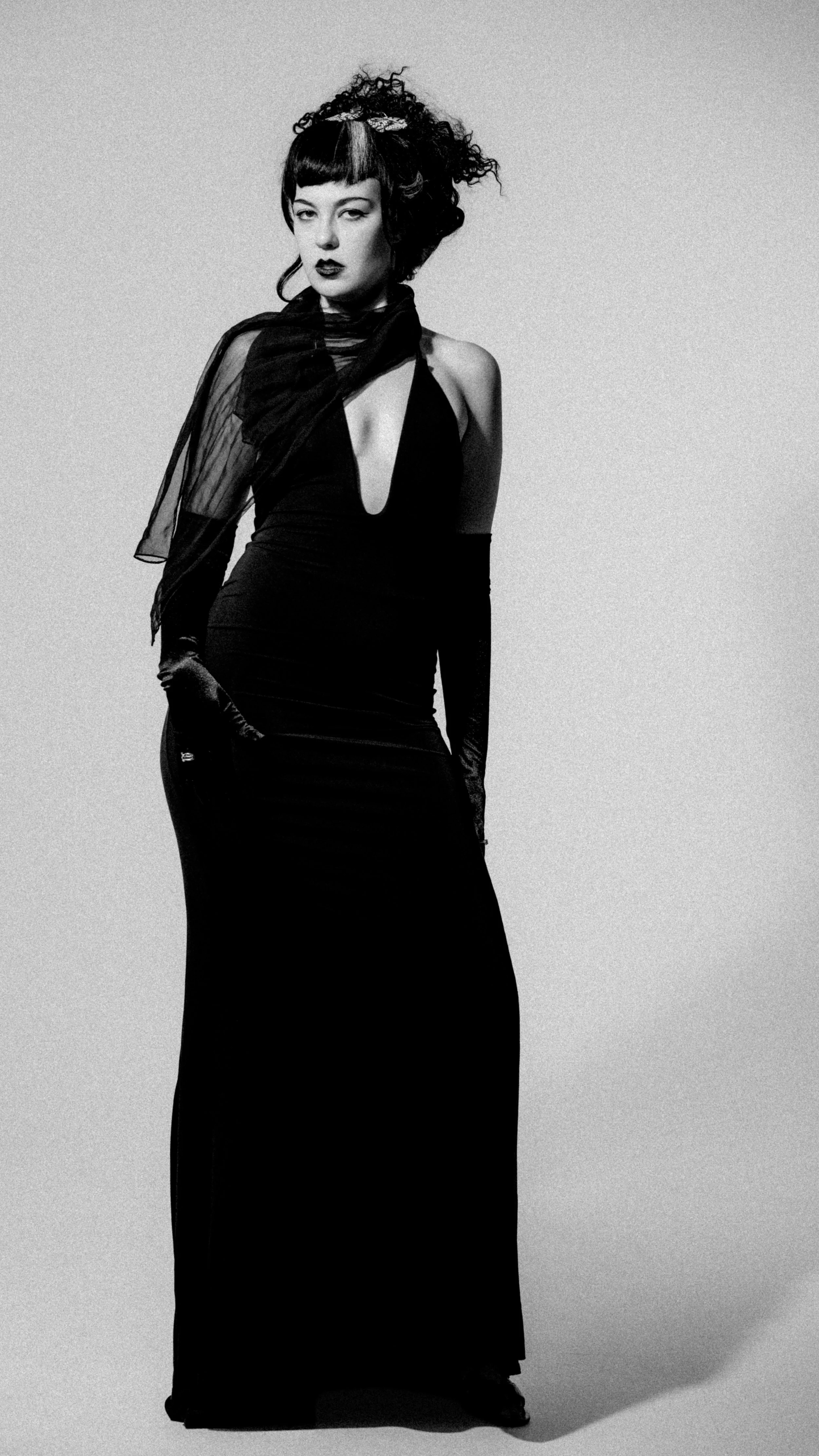 A black-and-white photo of a woman with short black hair, wearing a long black dress with a deep neckline, long gloves, and a sheer scarf. She has a confident expression, one hand on her hip, and stands against a plain background.