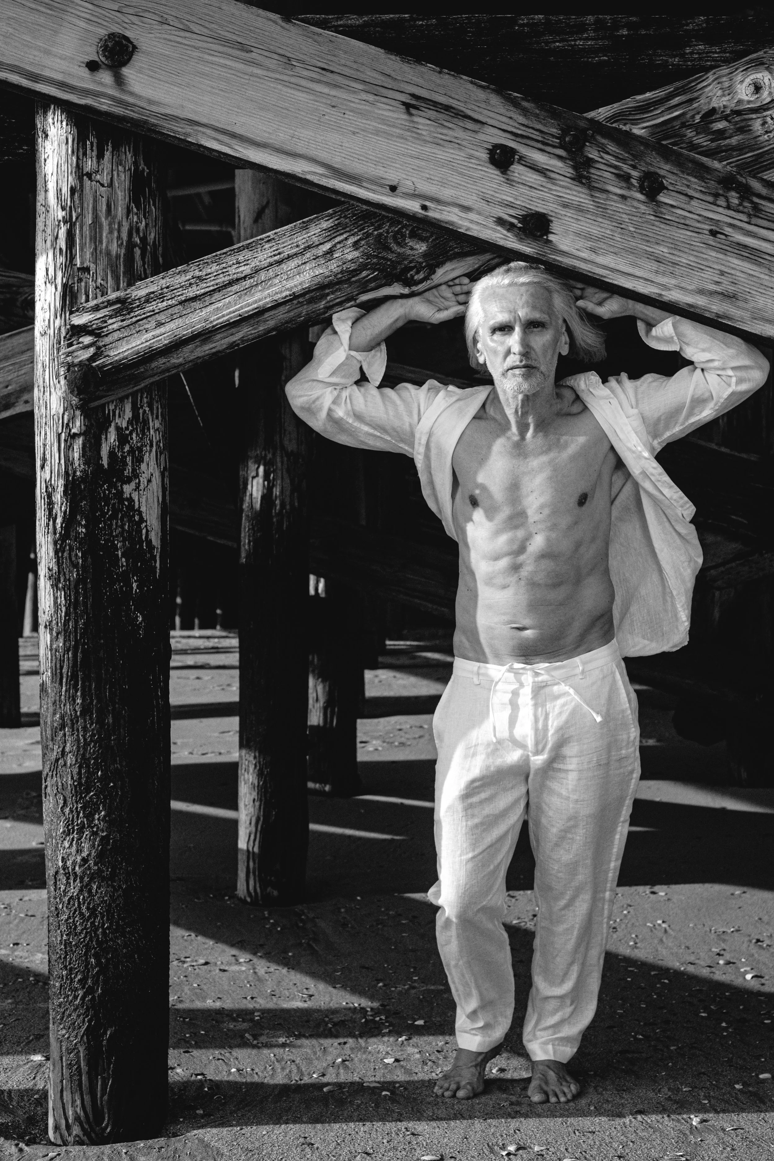 Black and white photo of a shirtless man with long hair standing barefoot on a sandy beach, holding a wooden beam on his shoulders, with wooden pilings and posts in the background.