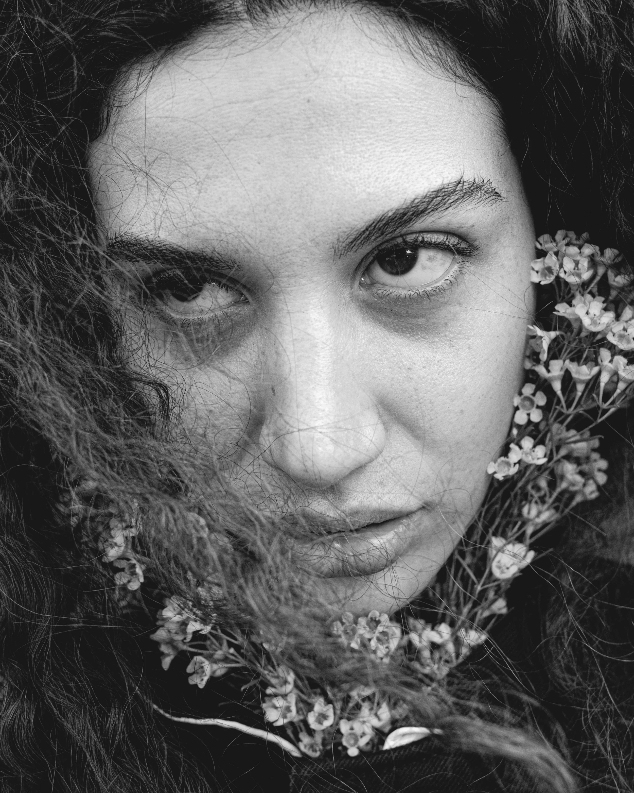 Close-up black and white photo of a woman's face, showing one eye and part of her nose, with her hair and small flowers around her face.
