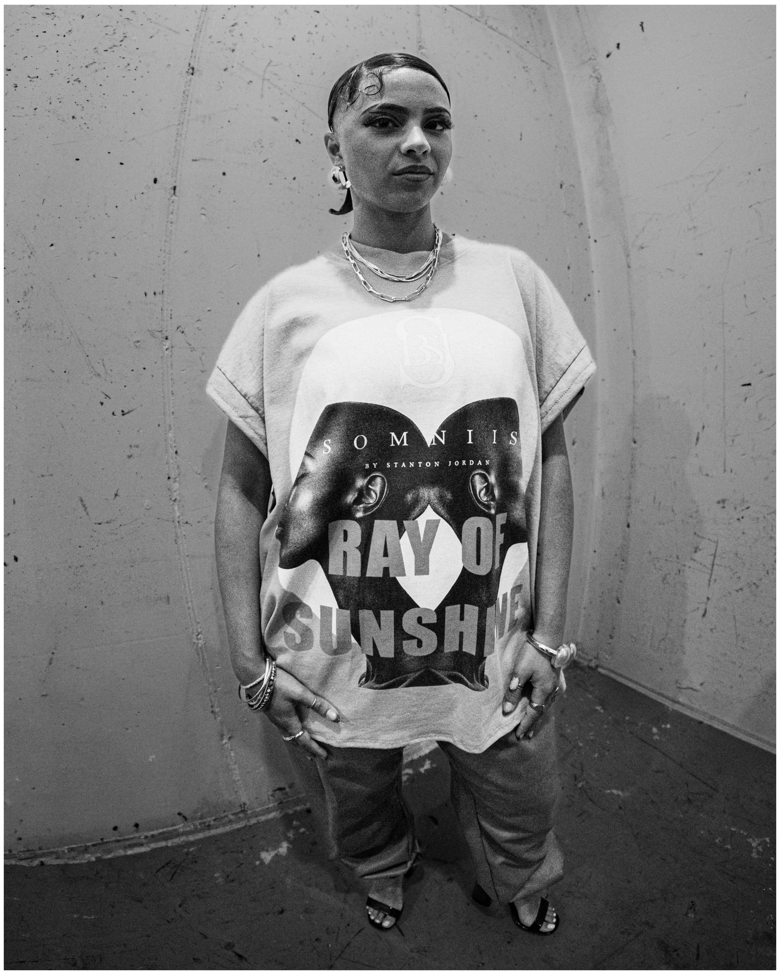 A person with short hair and earrings wearing a graphic t-shirt with text, multiple necklaces, and bracelets, standing against a textured wall.