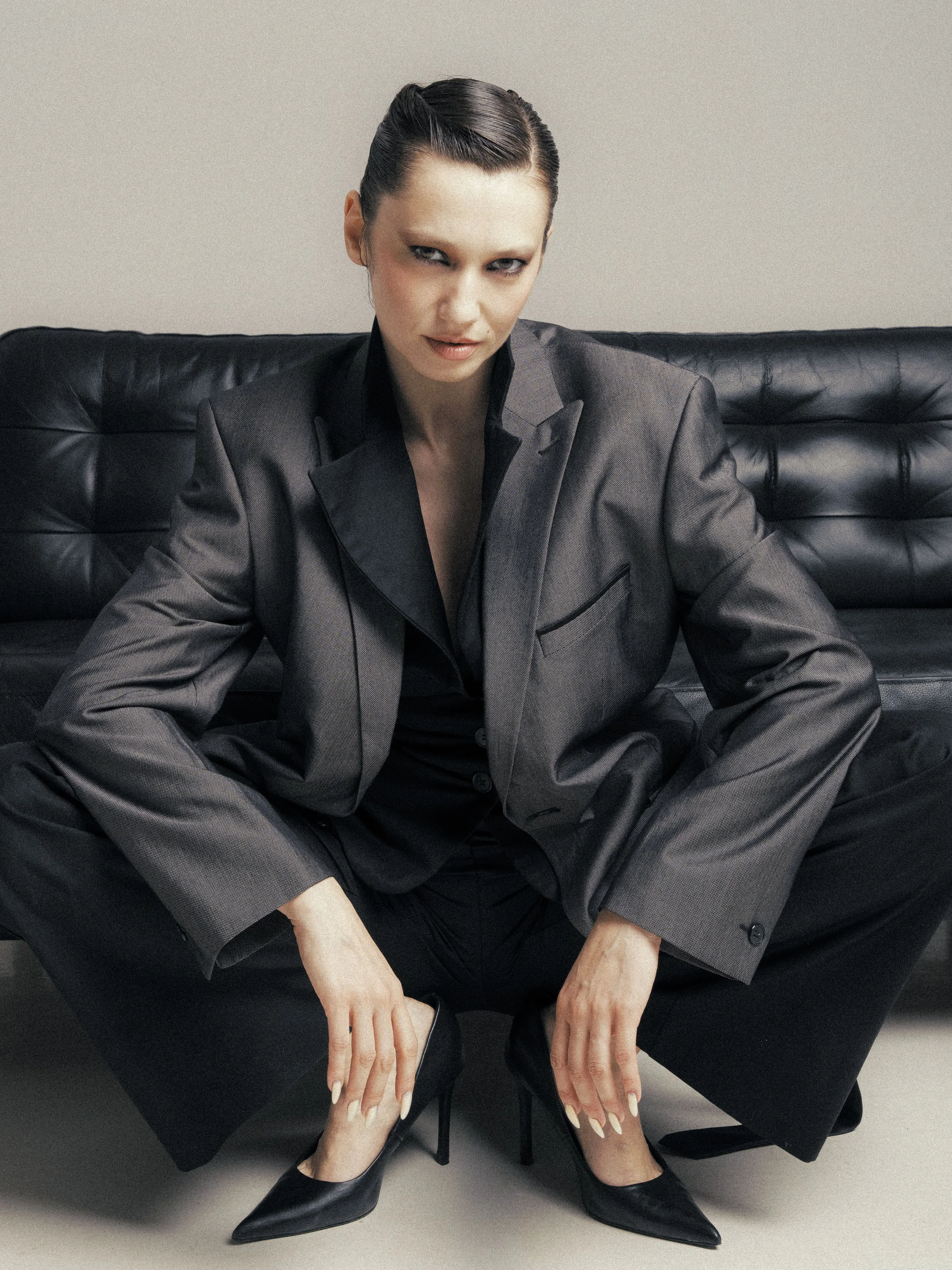 A woman with dark hair styled in a slicked-back fashion, wearing a dark gray suit, sitting with her legs apart on a black leather bench against a neutral background, looking directly at the camera with a confident expression.