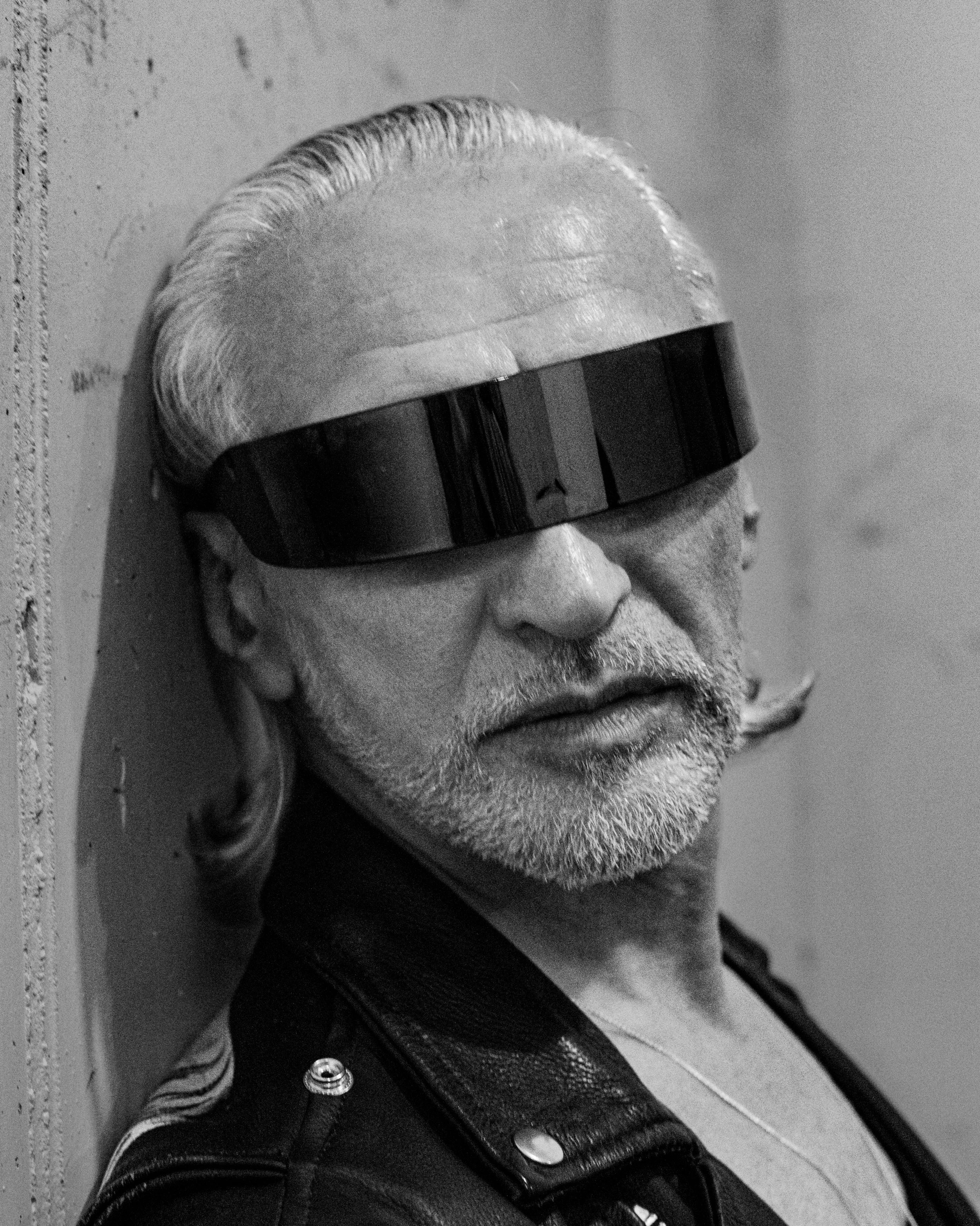 Close-up of a man's face with a beard and slicked-back hair, wearing large sunglasses, a leather jacket, and a striped shirt, lying on the ground next to a wall.