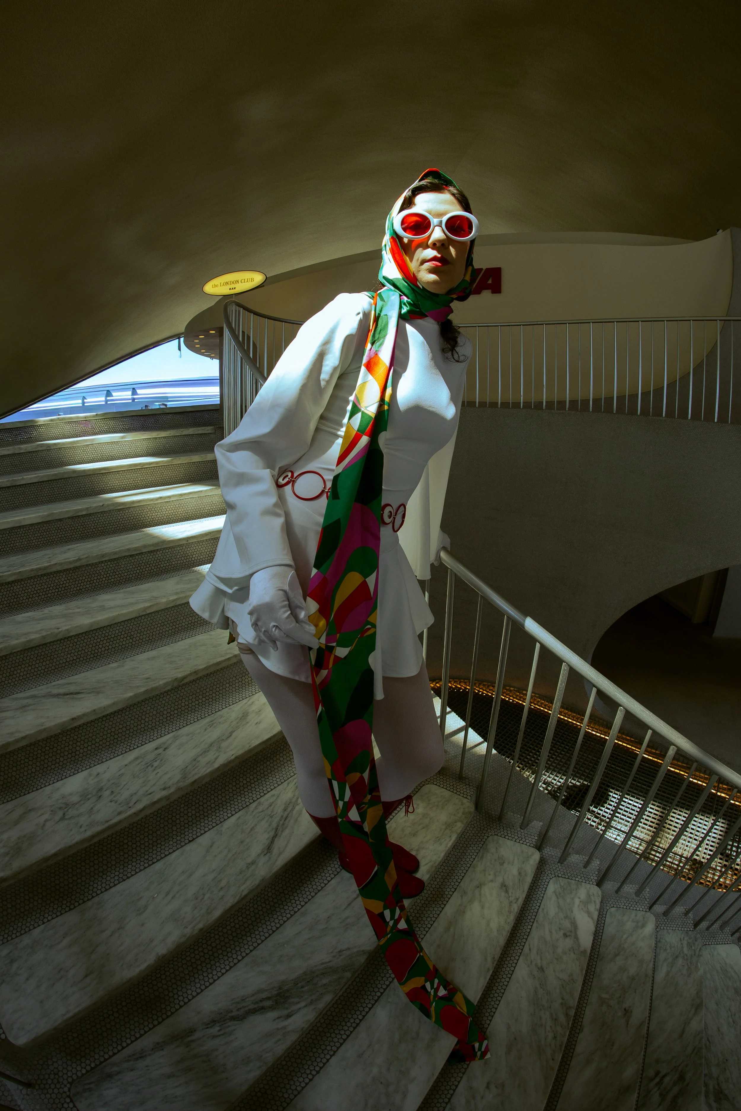 A woman dressed in all white with a colorful scarf, large red sunglasses, and red shoes standing on a marble staircase in an architectural setting.