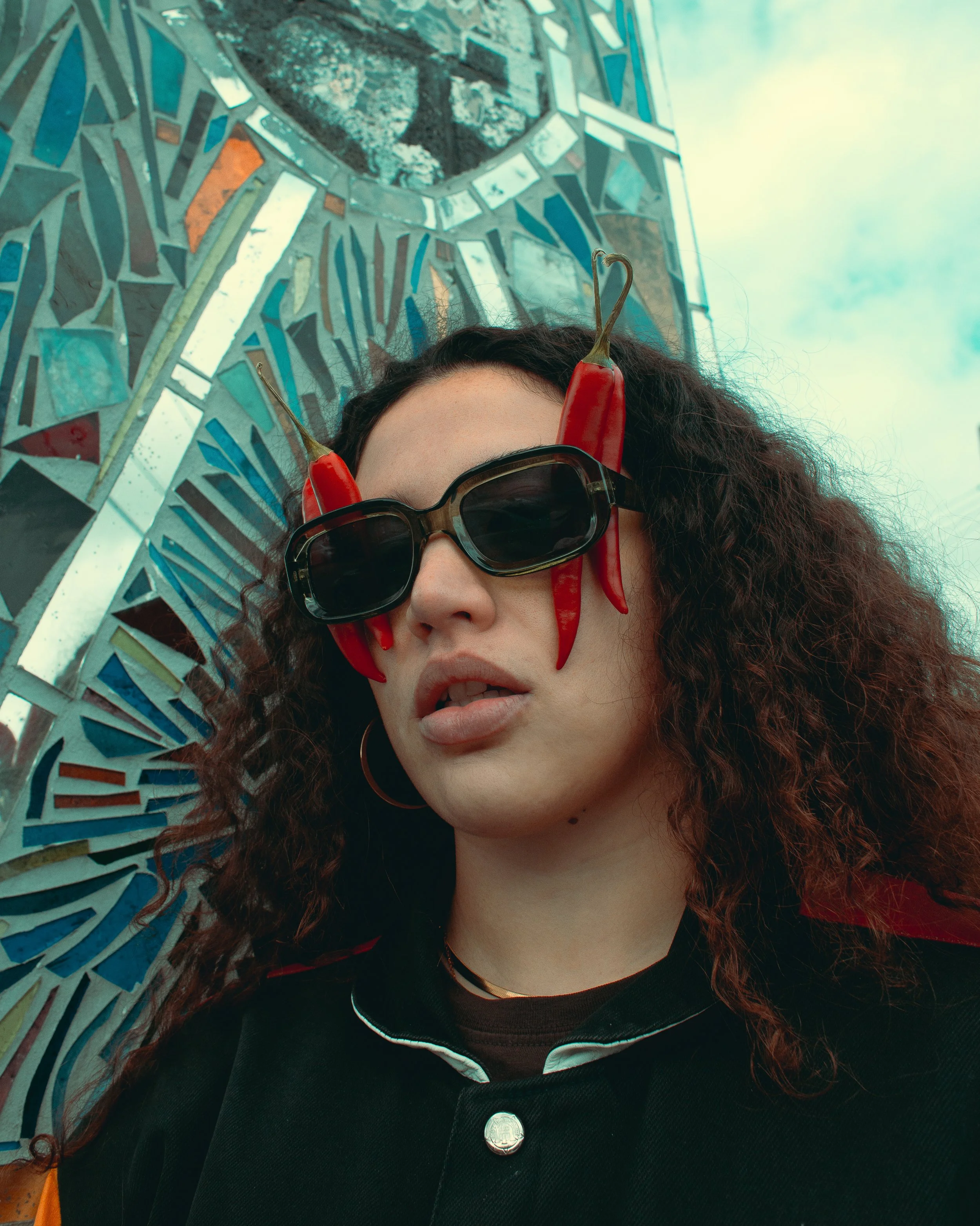 A woman with curly hair wearing sunglasses and a black jacket with a red chili pepper hanging on her glasses, standing in front of a mosaic mirror and an outdoor sky.
