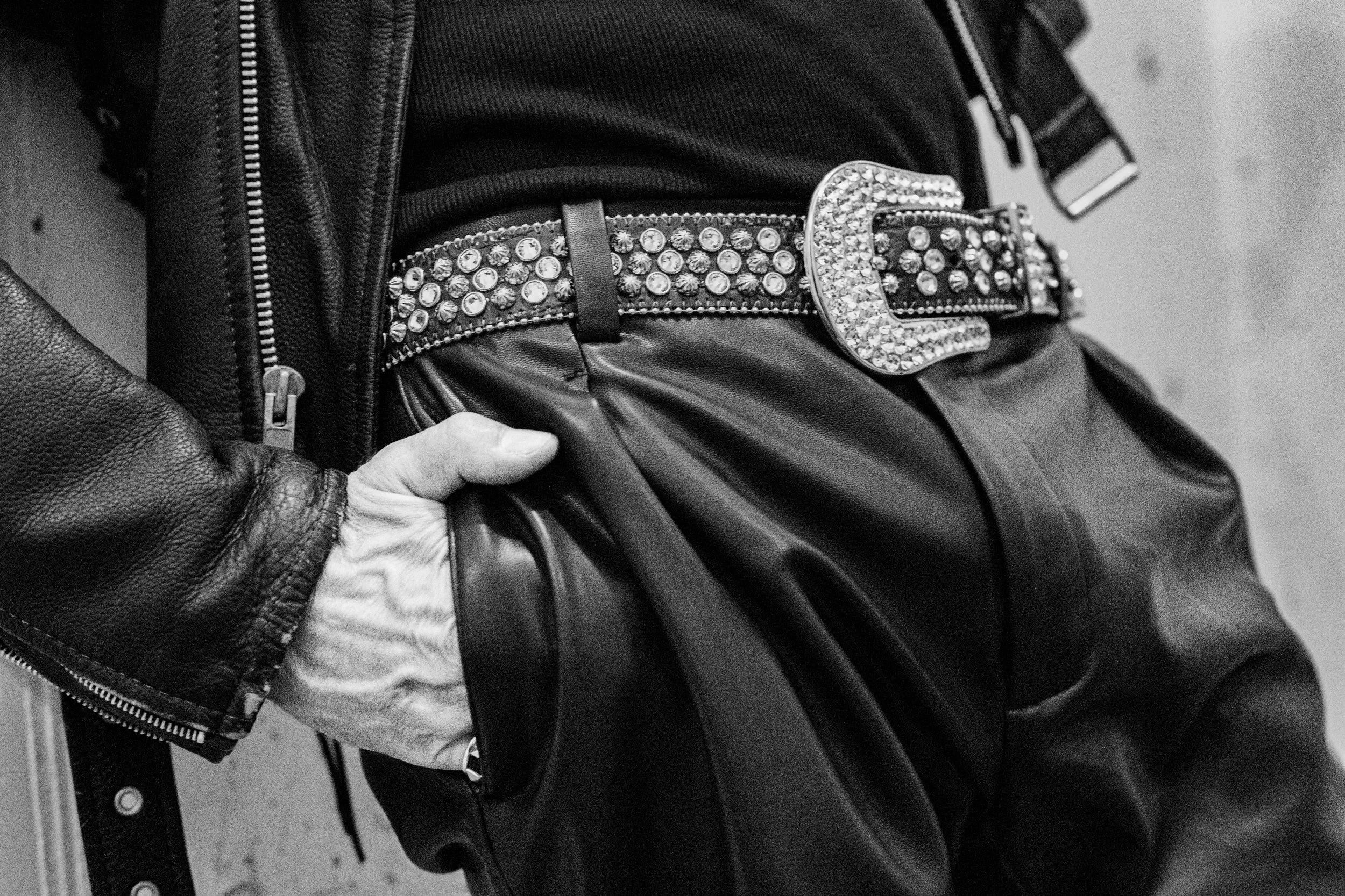 Close-up of a person's hand holding a black leather jacket with a zippered pocket, showing a wide belt with rhinestones and a large decorative buckle.