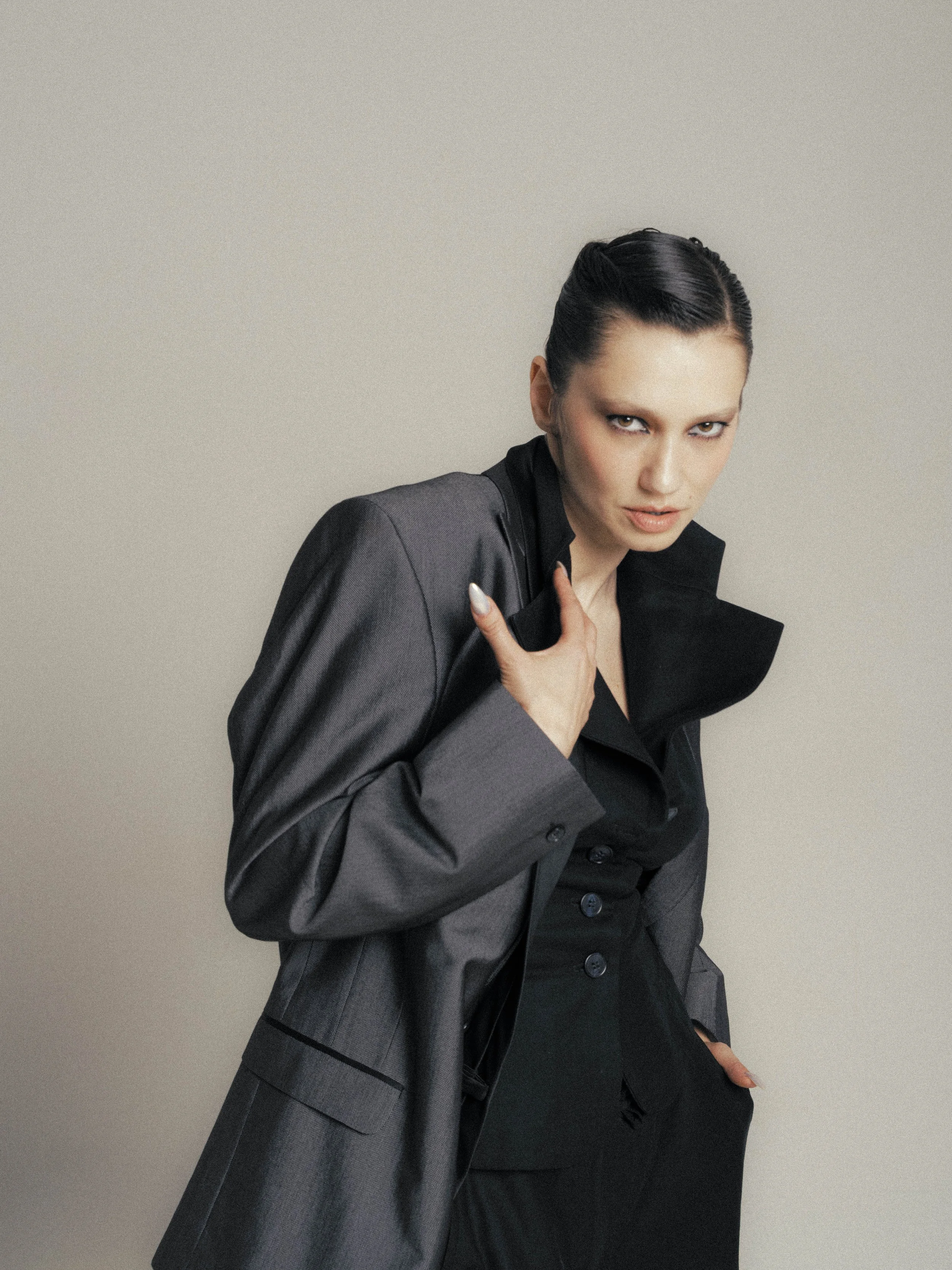 A woman with slicked-back black hair, wearing a black and gray suit, posing against a plain gray background.