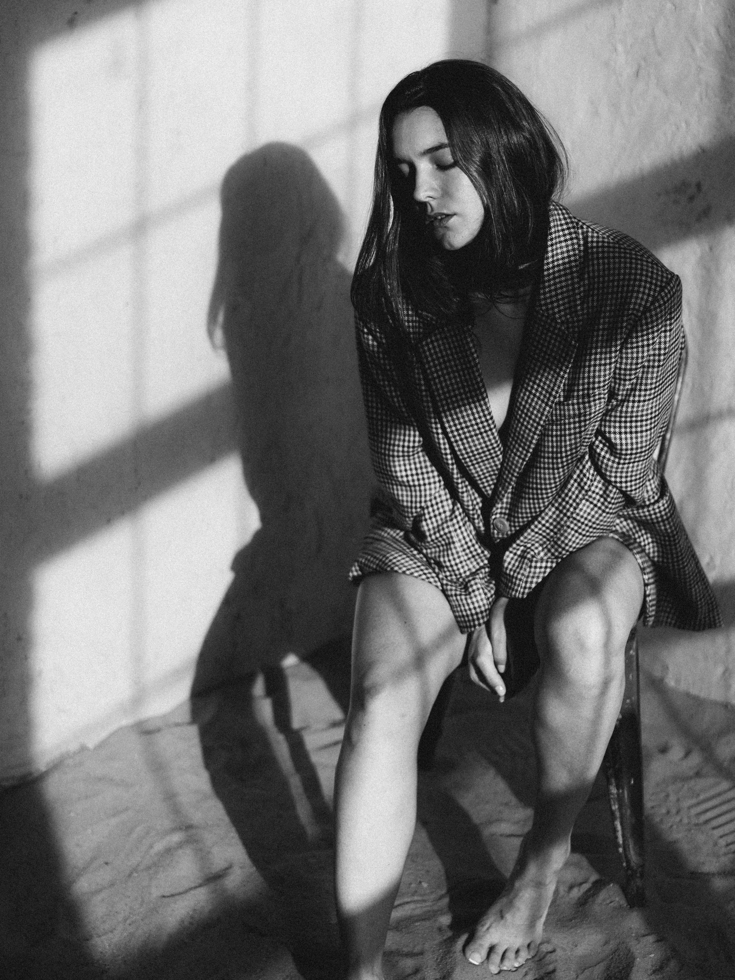 A black and white photo of a young woman sitting on a chair in a rustic setting. She has shoulder-length dark hair and is wearing a checked blazer with a low neckline. She is looking down with her eyes closed, with a contemplative expression on her f