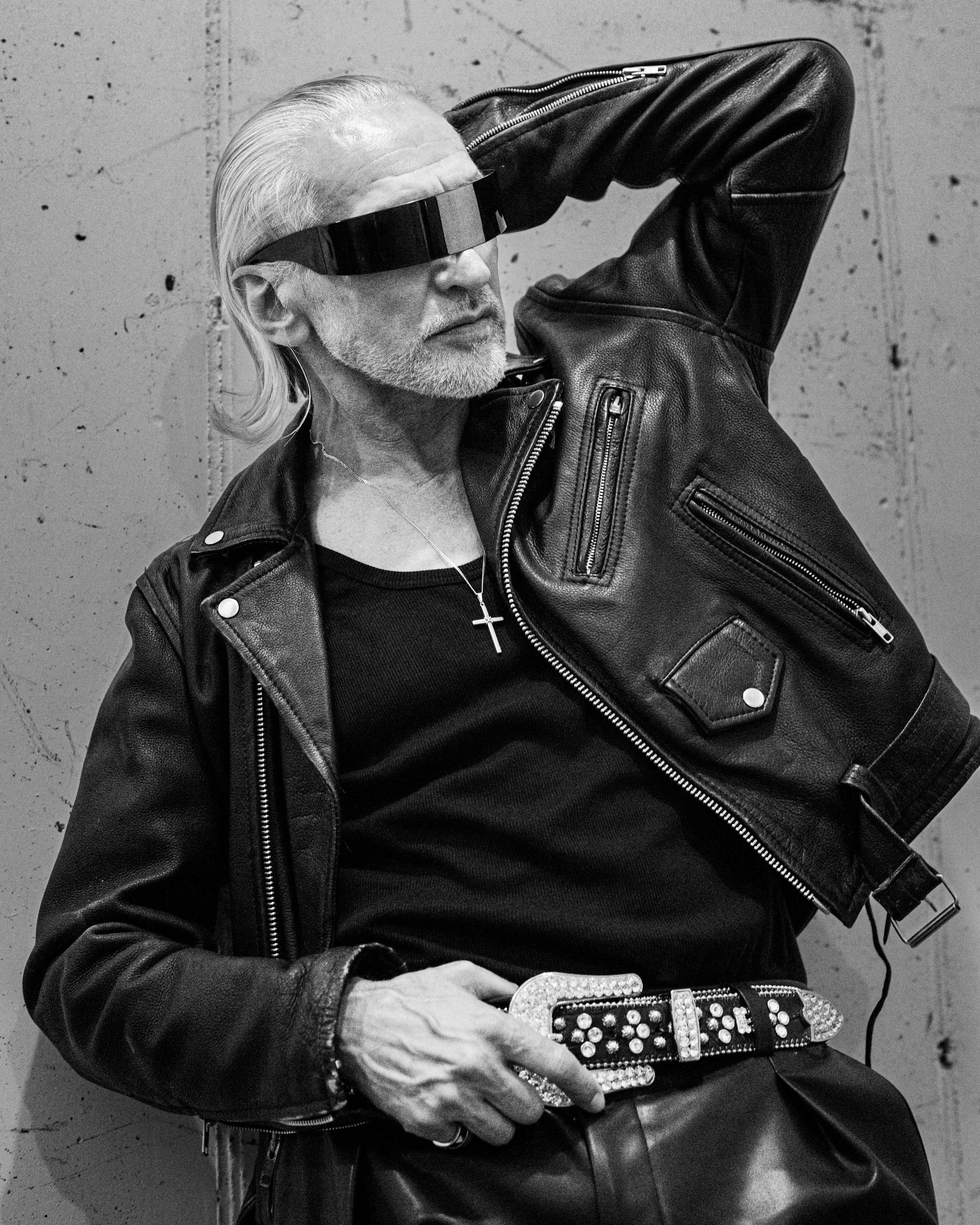 An older man with white hair and a beard is wearing a leather jacket and a visor glasses. He has a cross necklace and is holding a device with rhinestones, possibly a firearm or a jewelry item. The background is a plain, textured wall.