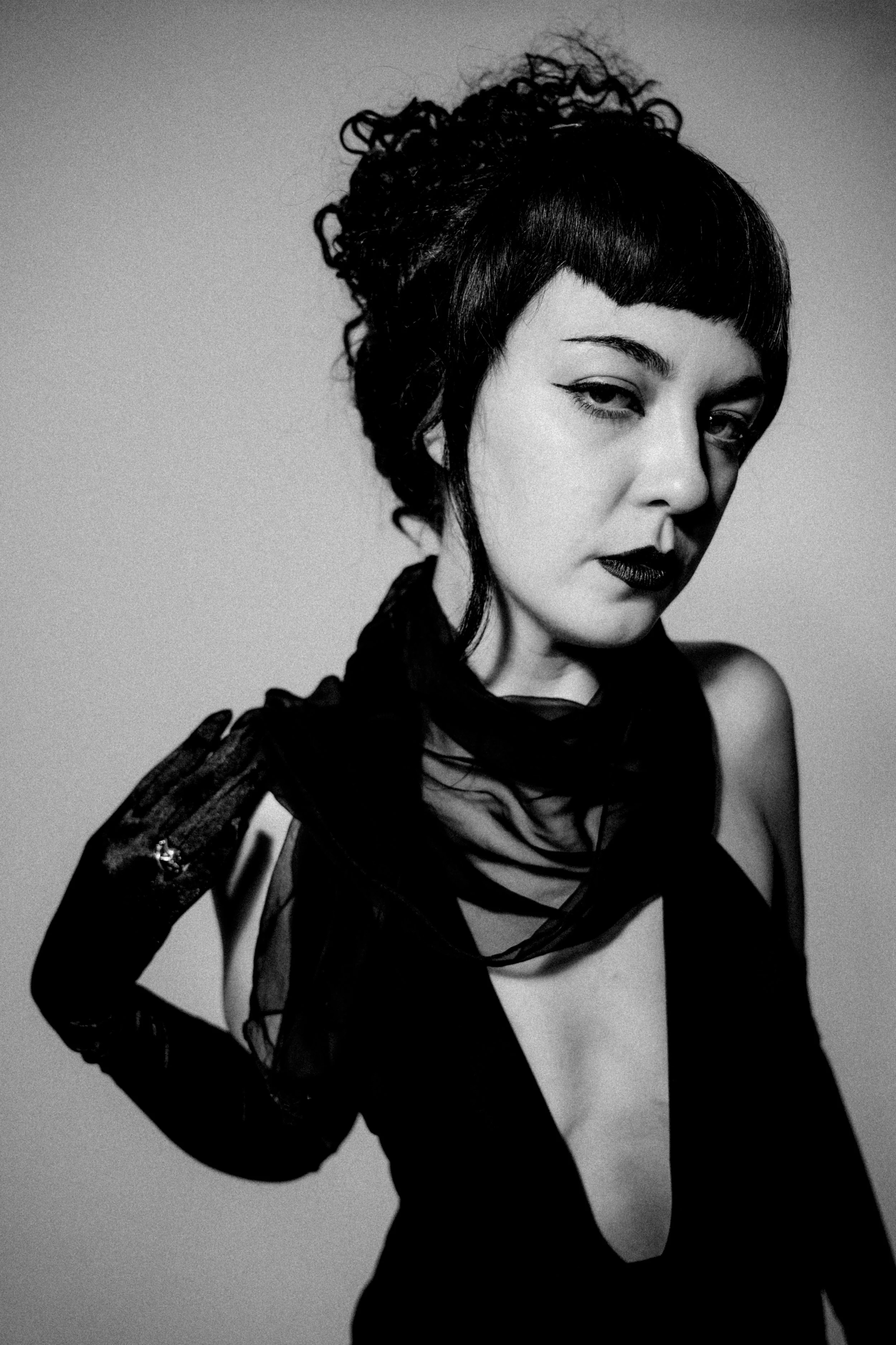 A woman with dark curly hair styled in a bun and blunt bangs, wearing a sheer black top with patterned long sleeves, tilts her head slightly with a confident expression.