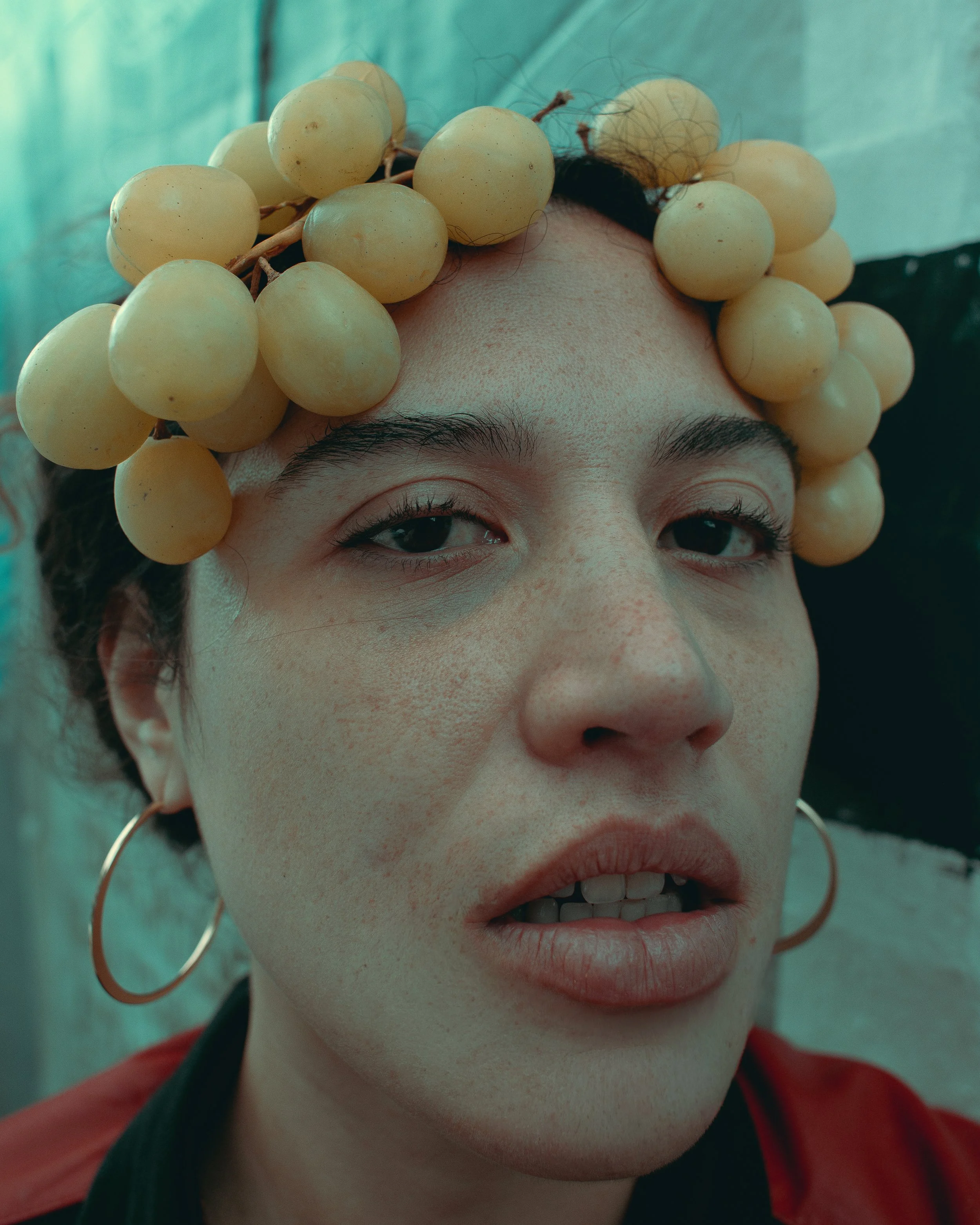 A person with a crown of white grapes on their head, wearing large gold hoop earrings, red lipstick, and a red and black outfit, smiling with slightly parted lips.