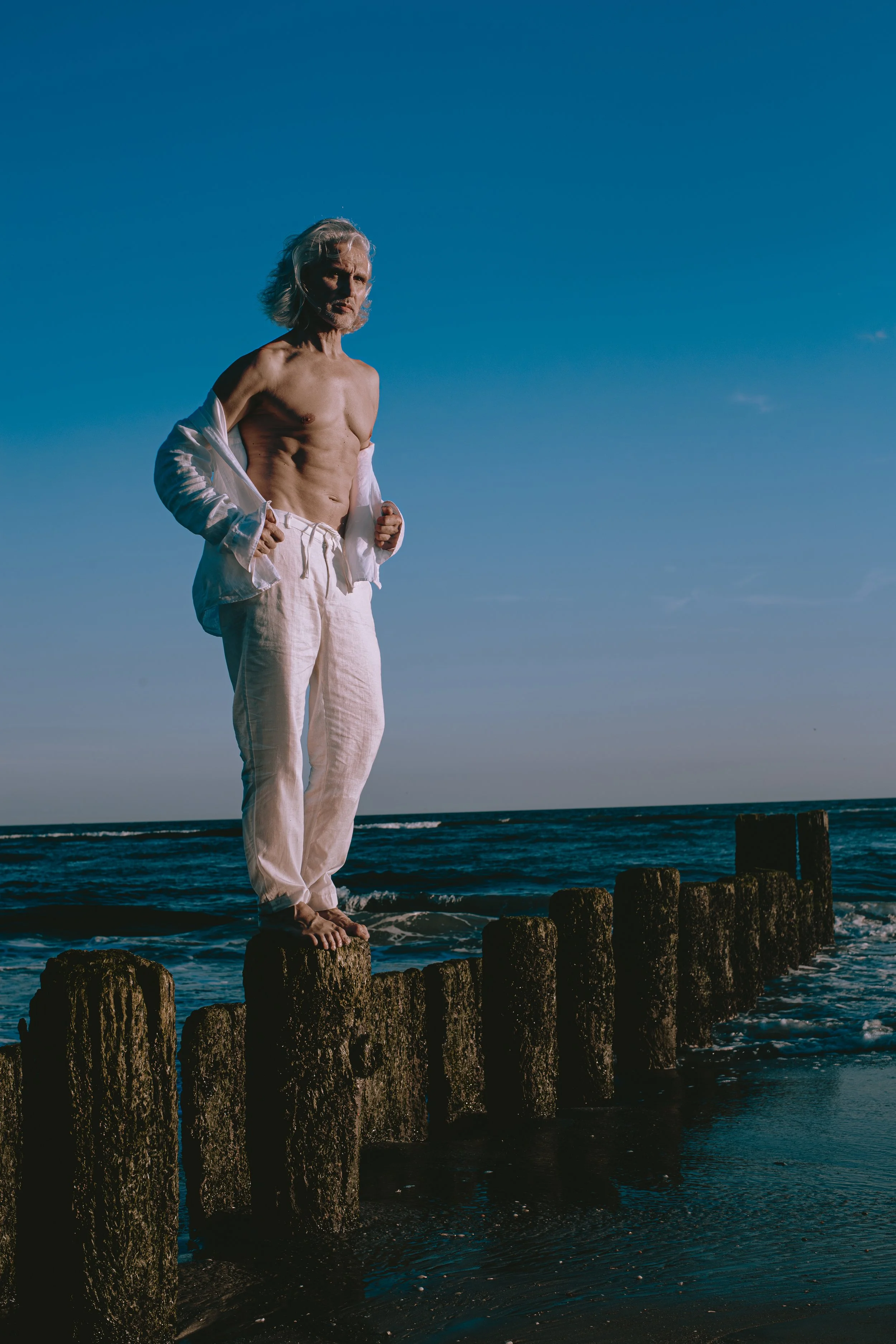 An elderly man with gray hair and a beard stands barefoot on wooden pilings by the ocean, shirtless and wearing white pants, with a white shirt unbuttoned and hanging off his shoulders, under a clear blue sky.