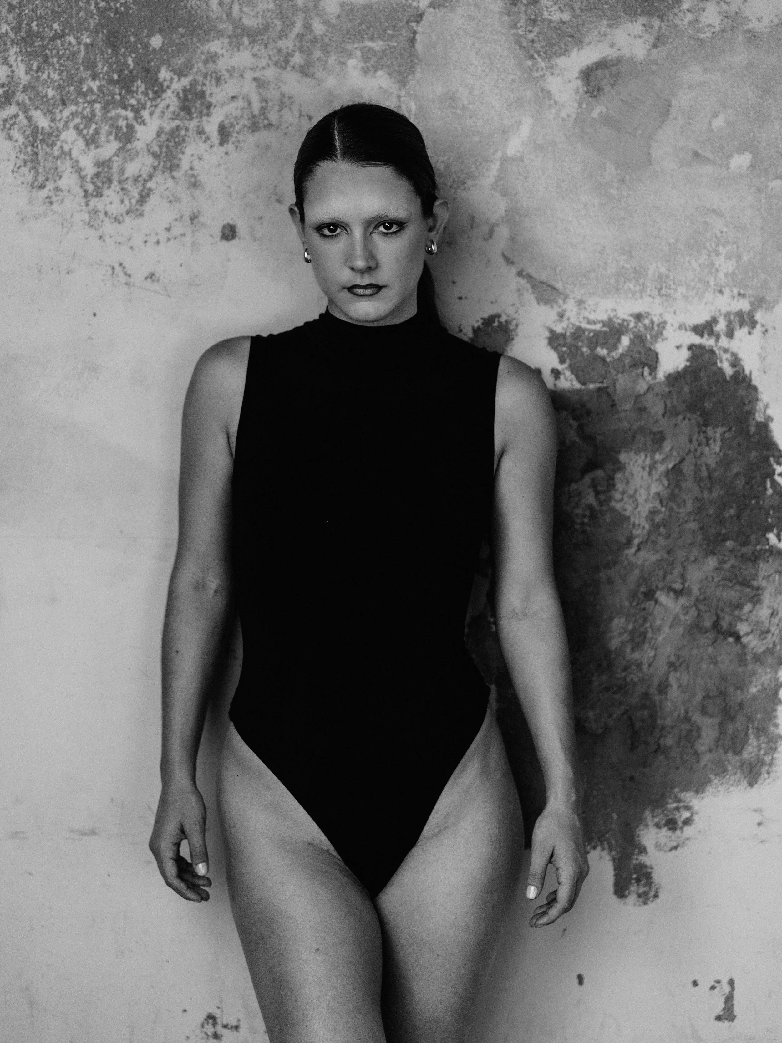 A black-and-white photo of a woman in a sleeveless black top and underwear, standing against a textured wall with peeling paint, looking directly at the camera.