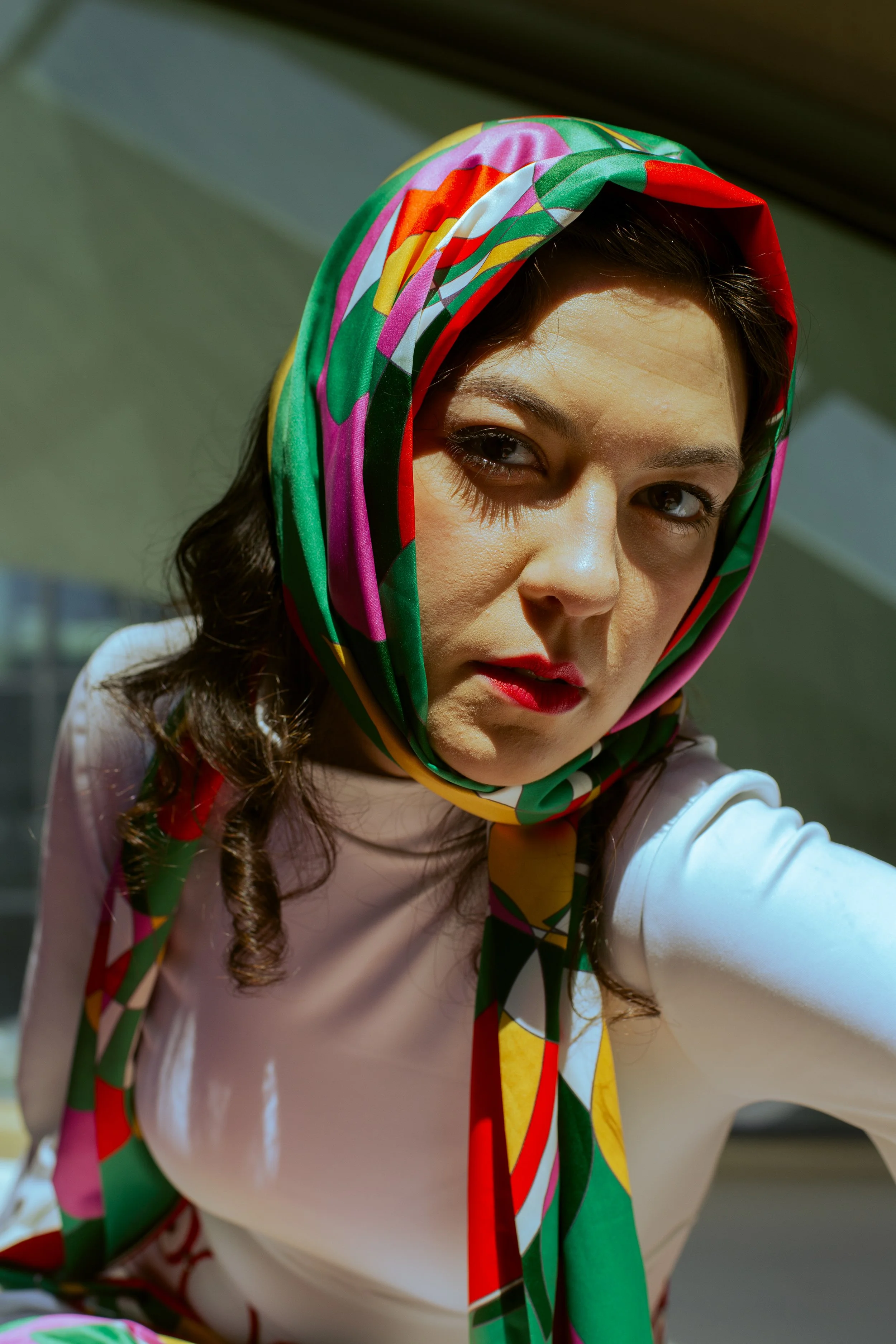 A woman with dark hair wearing a colorful, patterned scarf and a light-colored top, taking a selfie indoors near a window with sunlight illuminating her face.