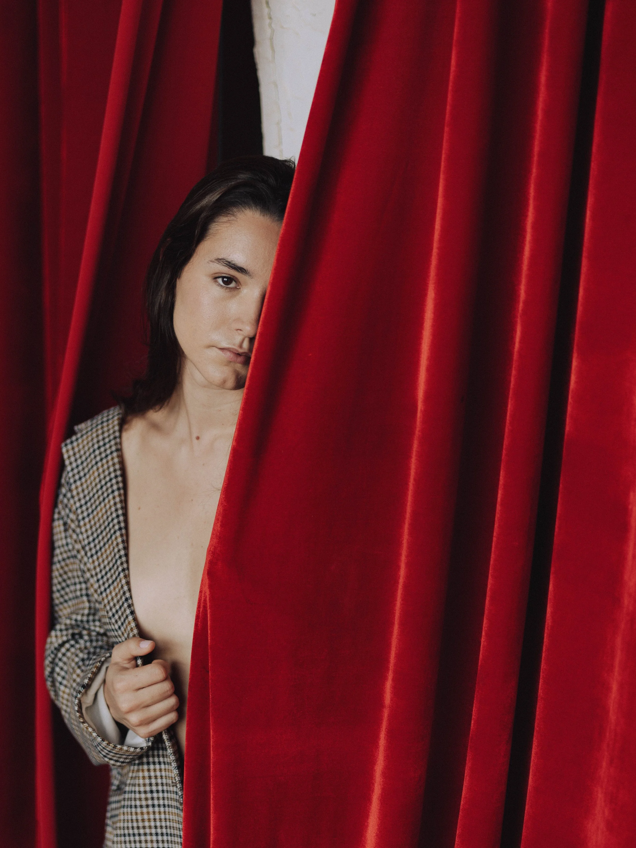 A woman with dark hair and light skin peeking out from behind a red theatre curtain, holding it with her hand.
