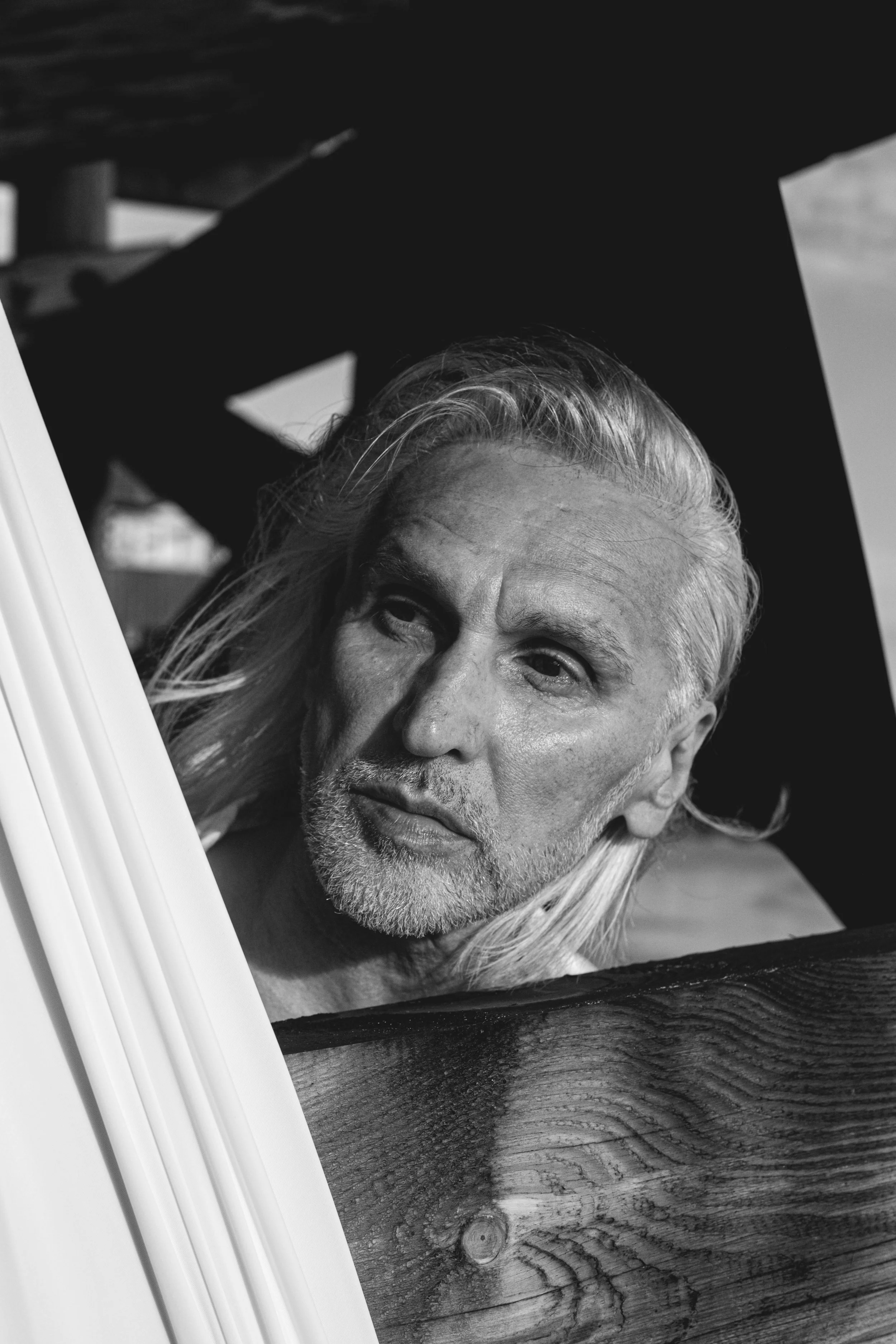 A black and white close-up photo of a middle-aged man with long, wavy hair and a beard, looking into a mirror.