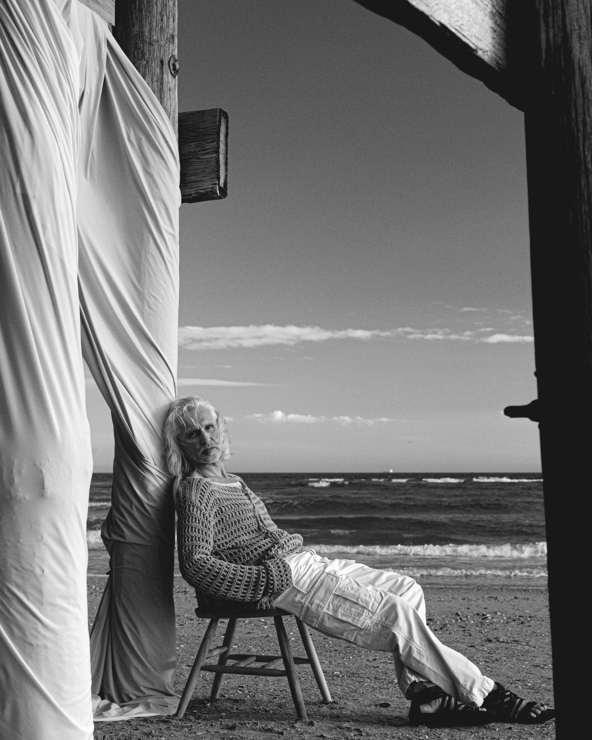 A man with long hair and a beard sitting on a wooden chair on the beach, gazing at the camera; a woman standing next to him with her face turned away, wearing a long skirt. The scene is set against the ocean and a partly cloudy sky.